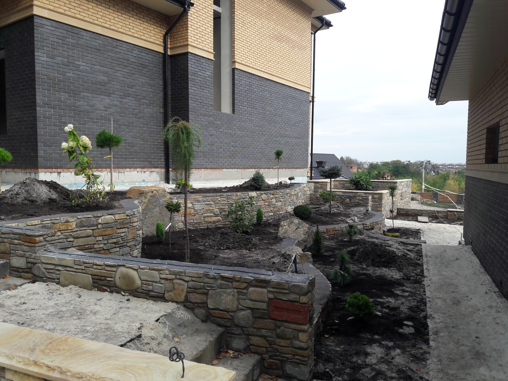 Ogród z kamiennymi murkami kaskadowymi i świeżo posadzoną roślinnością, na tle dwukondygnacyjnego domu z ciemnej cegły klinkierowej i jasną elewacją na górze.