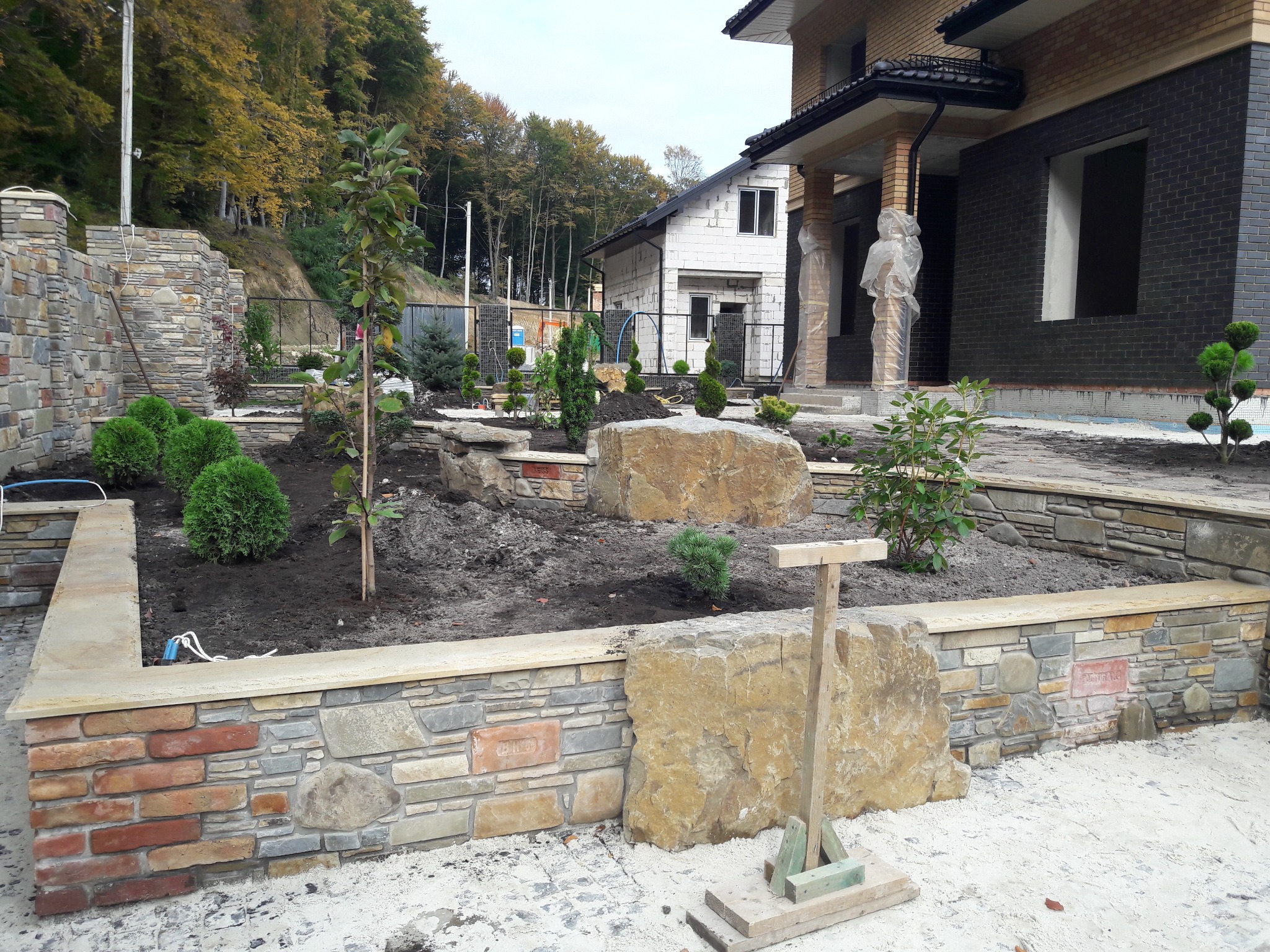 Ogród w trakcie aranżacji: kamienne murki, nasadzenia drzew i krzewów, w tle dom z elewacją z ciemnej cegły i drugi budynek w stanie surowym. Widoczne elementy konstrukcyjne.