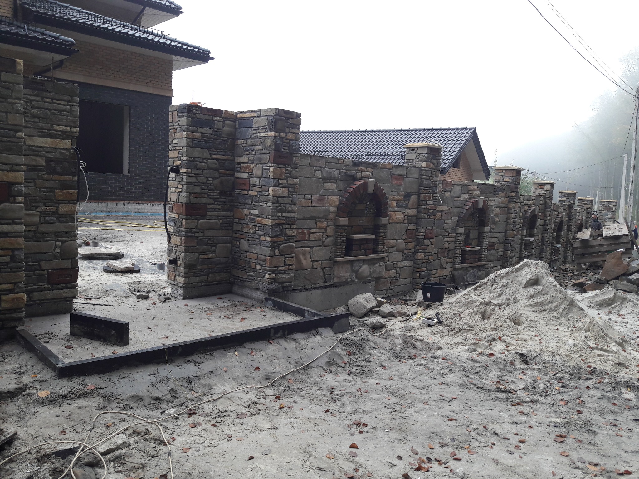 Wznoszony kamienny mur z łukowymi wnękami, fragment domu w tle. Widoczne materiały budowlane i elementy konstrukcyjne. Etap budowy w pochmurny dzień.