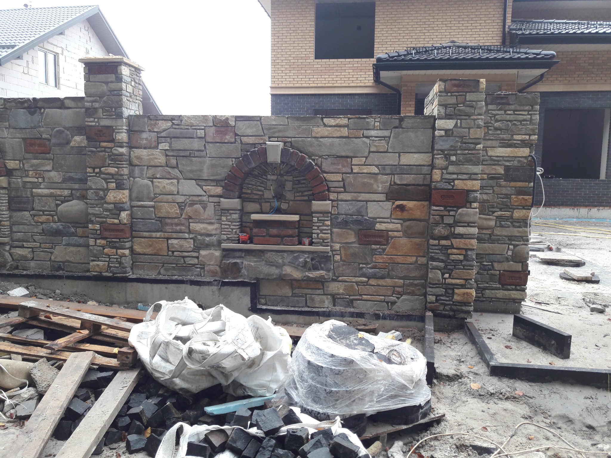 Kamienny mur z łukiem i kolumnami, fragment elewacji domu w tle. Widoczne materiały budowlane i elementy wykończeniowe. Ujęcie z bliska, podkreślające fakturę kamienia.