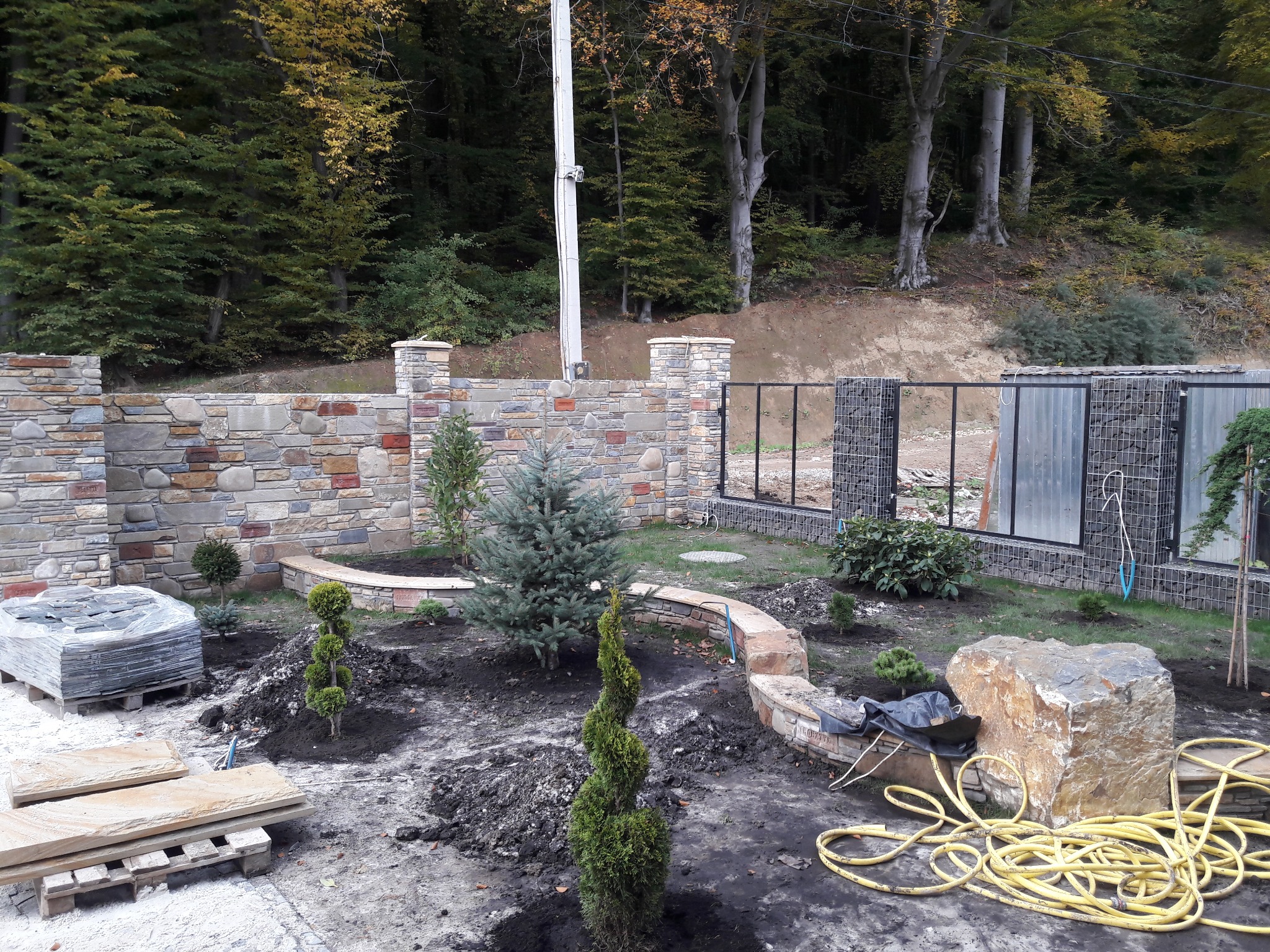 Aranżacja ogrodu w trakcie realizacji: kamienne murki, gabiony, przygotowane sadzonki drzew i krzewów, kamienne płyty tarasowe. Widok na zalesione wzgórze.