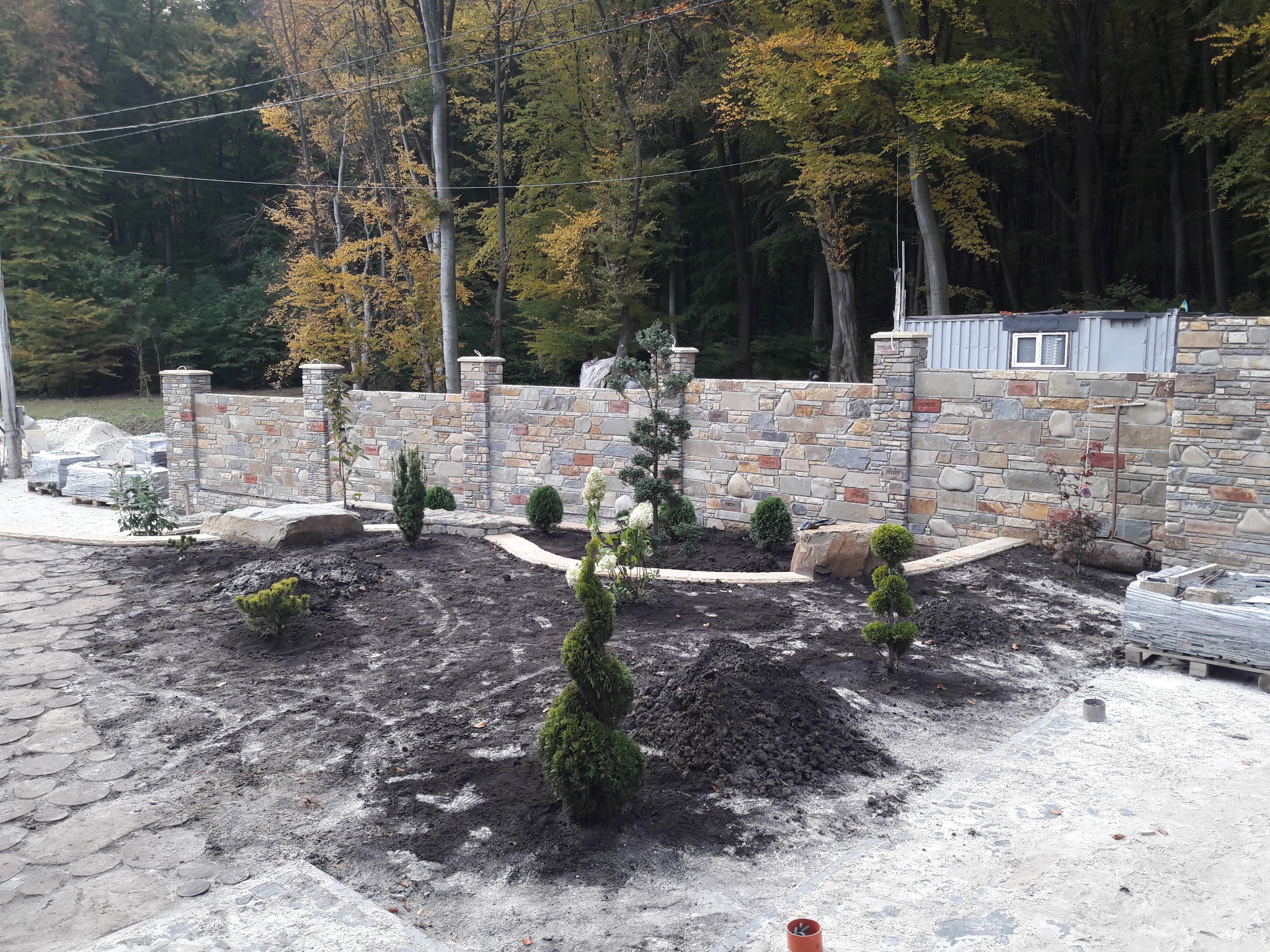 Kamienny mur z kolumnami i nowo posadzone rośliny ozdobne na tle lasu. Widoczny fragment kamiennego chodnika i paleta z płytami.