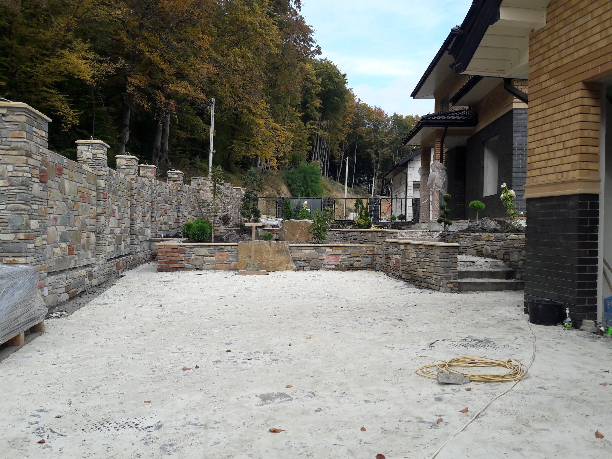 Kamienny taras z niskim murem i kolumnami z kamienia, w tle dom z elewacją z cegły klinkierowej i drzewa. Widoczne materiały budowlane na palecie.