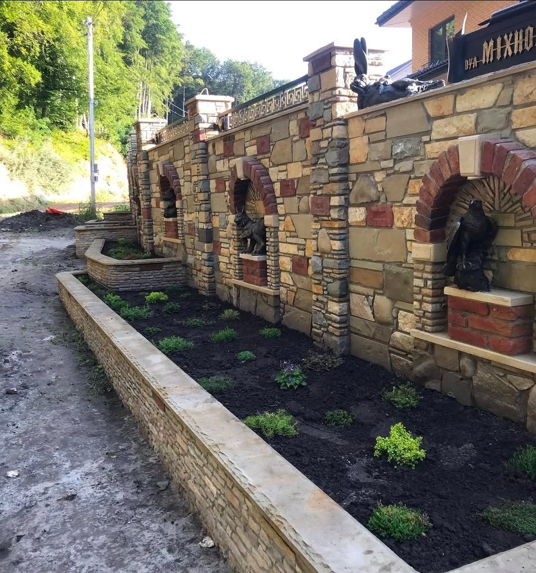 Kamienny mur z dekoracyjnymi wnękami i rzeźbami zwierząt, oddzielający posesję od drogi. Wzdłuż muru rabata z nasadzeniami. Widoczne elementy wykończenia z cegły.