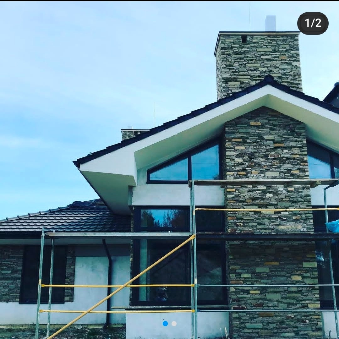 Elewacja domu w trakcie prac: kamienna okładzina na ścianie i kominie, widoczne rusztowania. Nowoczesna architektura z dużymi oknami i skośnym dachem.
