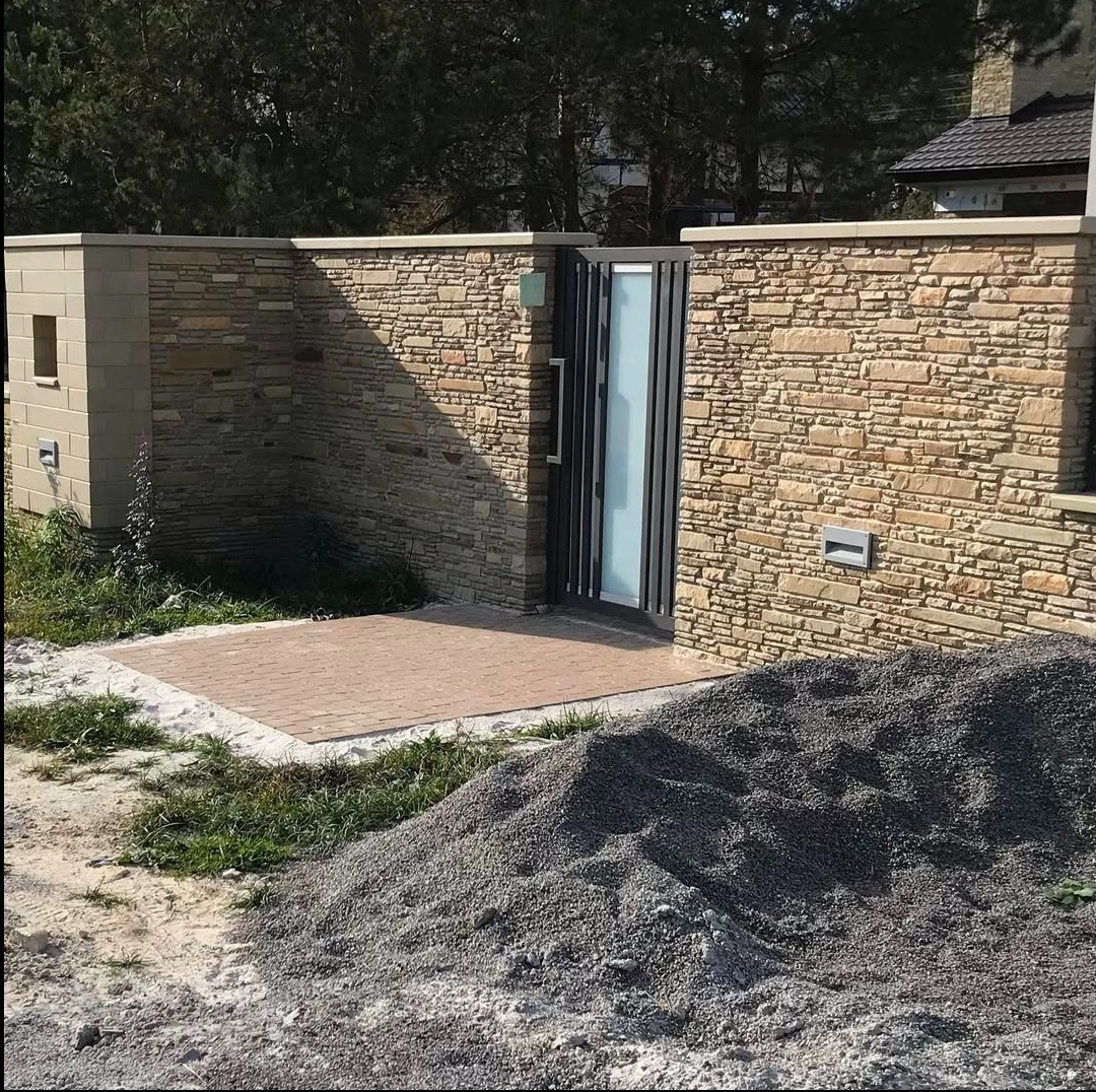 Kamienny mur z furtką i oświetleniem zewnętrznym, wykończony kostką brukową. W tle widoczny dom i drzewa. Na pierwszym planie hałda żwiru.