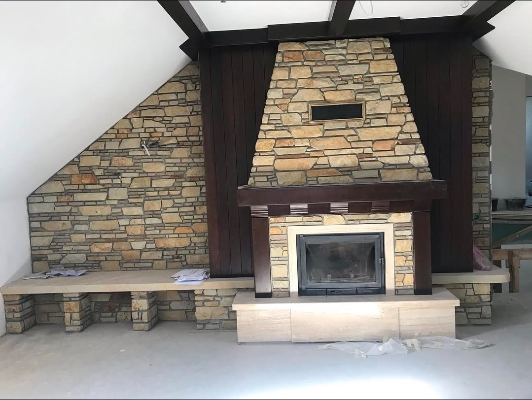 Kamienny kominek z drewnianą obudową i siedziskiem, wykończony w jasnych barwach, z widoczną fakturą kamienia i drewnianymi elementami dekoracyjnymi.