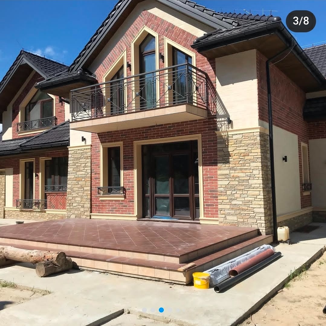 Dwukondygnacyjny dom z cegły klinkierowej z balkonem i tarasem wyłożonym płytkami. Widoczne detale architektoniczne i elementy wykończeniowe.