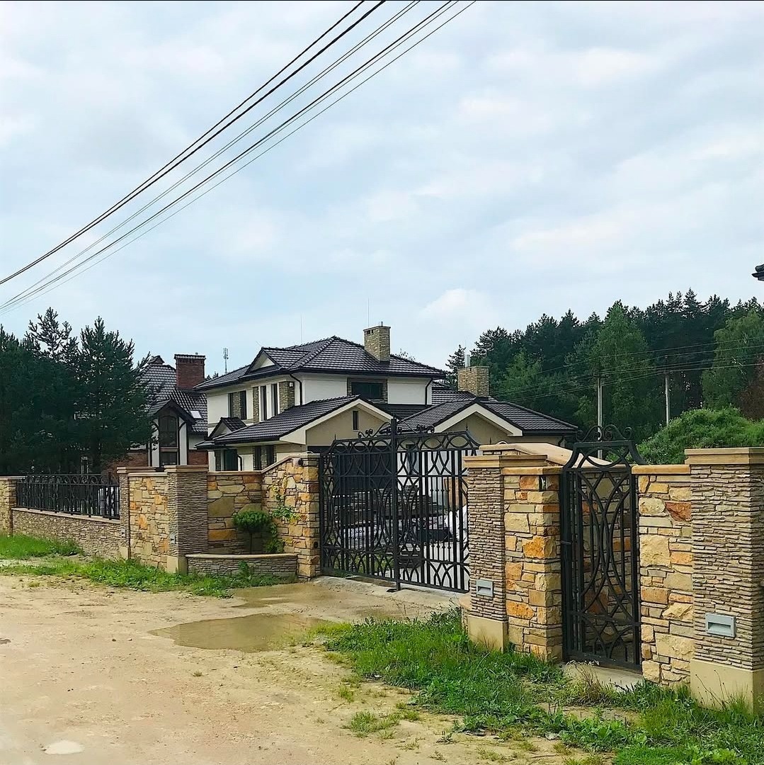 Nowoczesny dom z ciemnym dachem i kamiennym ogrodzeniem z kutą bramą. Widok z zewnątrz, pochmurne niebo w tle.