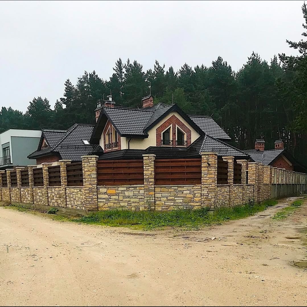 Nowoczesny dom z ciemną dachówką i kamienno-drewnianym ogrodzeniem na tle lasu. Ujęcie z poziomu gruntu, podkreślające detale elewacji i ogrodzenia.
