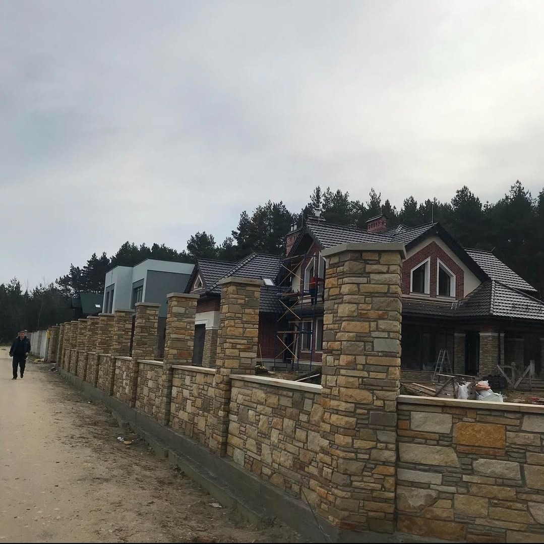 Wznoszony dom z cegły z wykończeniem z kamienia, widoczny zza kamiennego ogrodzenia. Na rusztowaniu pracownik. W tle las i pochmurne niebo.