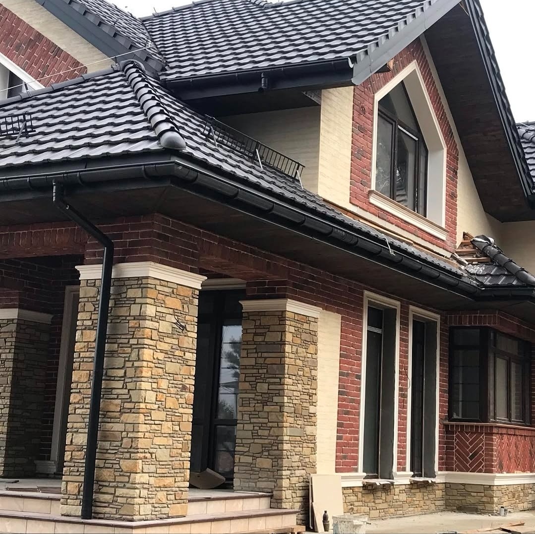 Dwukondygnacyjny dom z elewacją z czerwonej cegły i kamienia, czarną dachówką i oknami z białymi ramami. Widoczne detale wykończeniowe i elementy dekoracyjne.