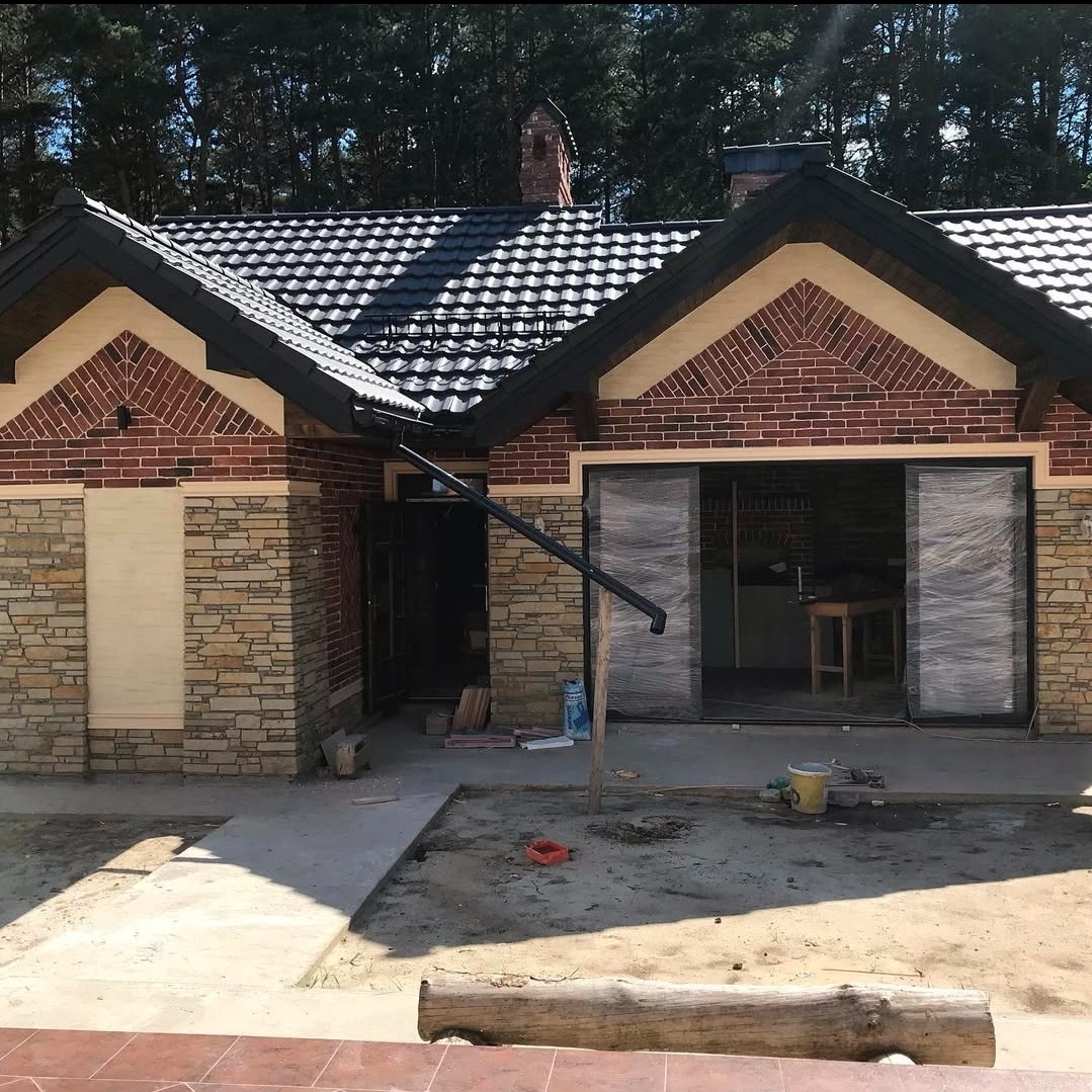 Dwukondygnacyjny dom z elewacją z kamienia i cegły, czarnym dachem i dużymi przesuwnymi drzwiami, częściowo owiniętymi folią ochronną. Widoczne narzędzia i materiały budowlane.