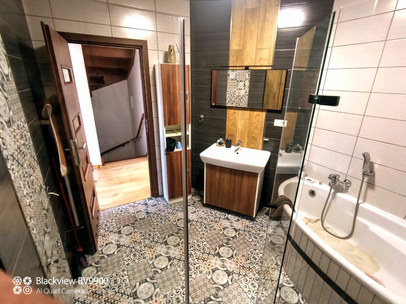 Nowoczesna łazienka z wzorzystą podłogą, drewnianymi akcentami na ścianach i szafce pod umywalką, oraz widoczną kabiną prysznicową i otwartymi drzwiami do klatki schodowej.