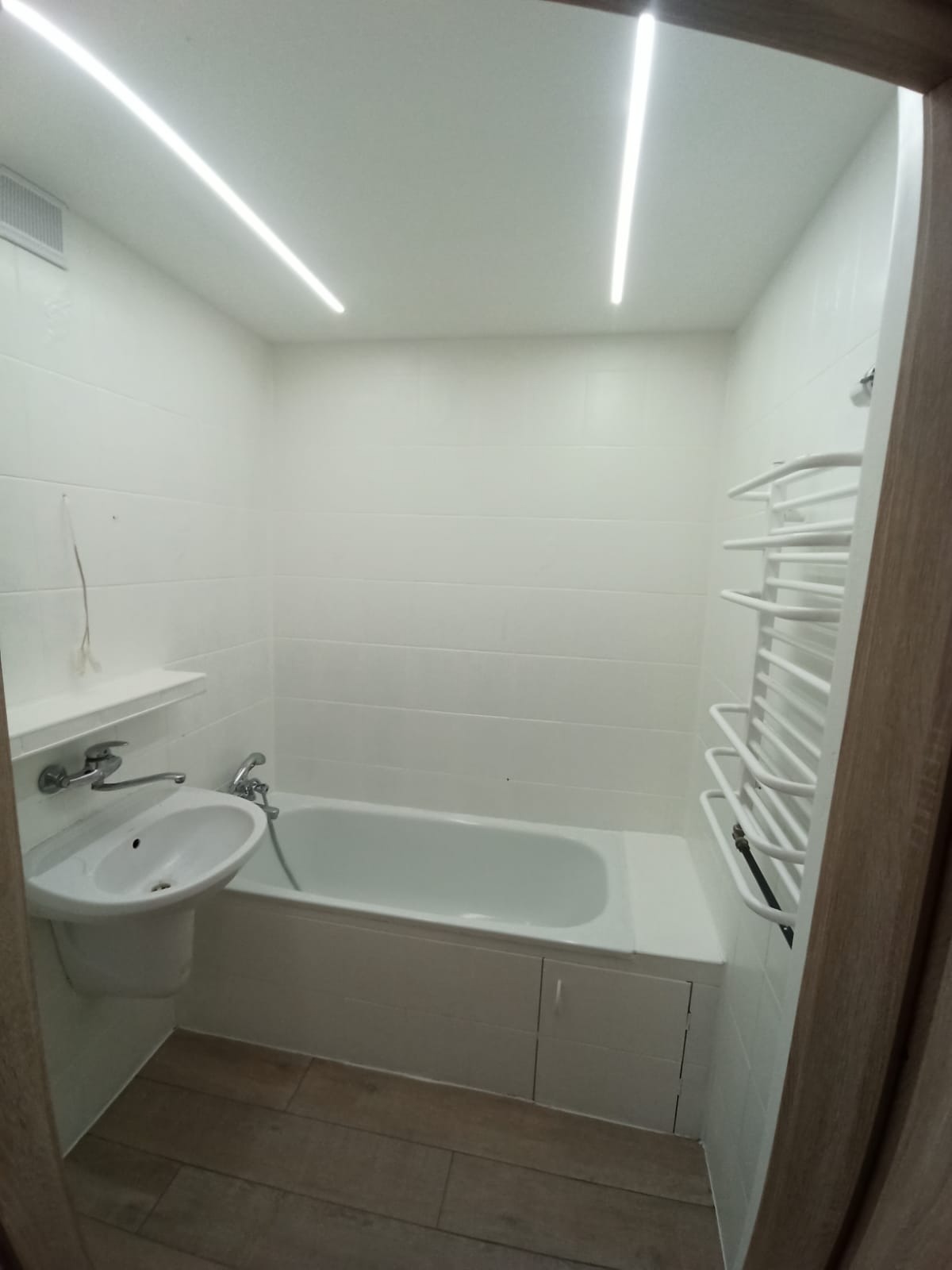 Jasna łazienka z białą armaturą, grzejnikiem drabinkowym i podłużnym oświetleniem LED podsufitowym, widok zza framugi drzwi.