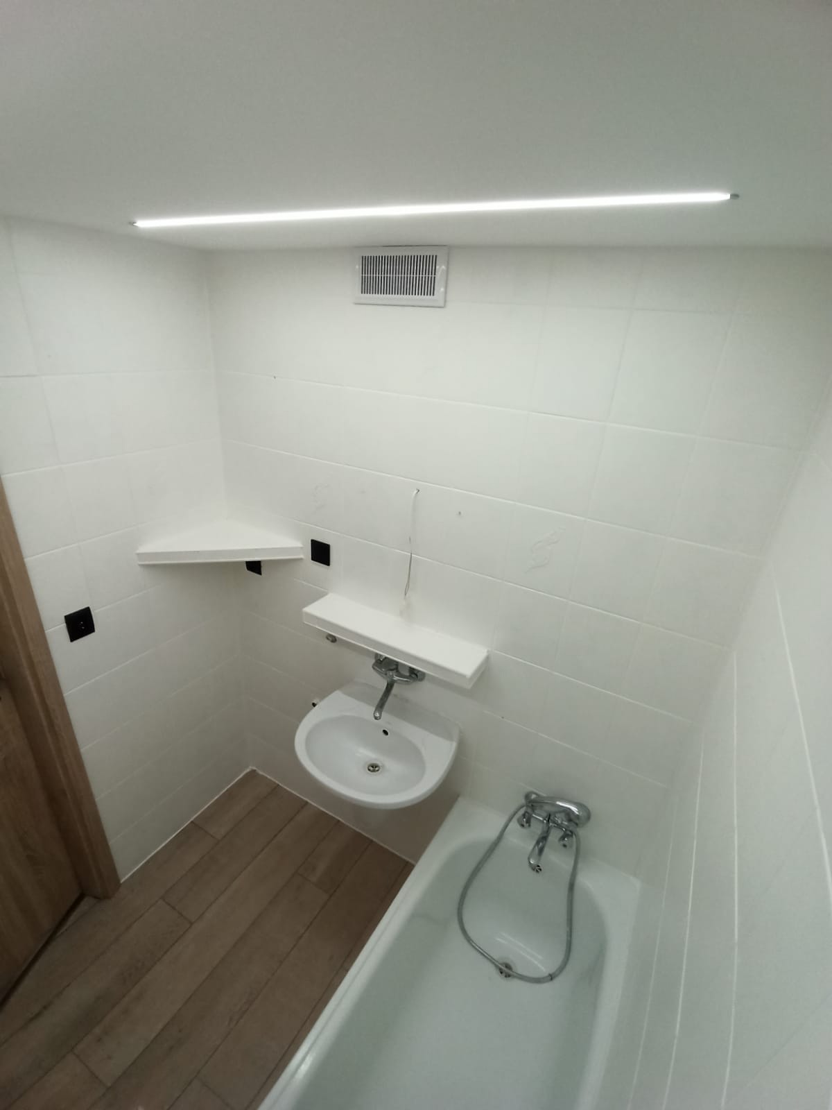 Mała, odnowiona łazienka z białą glazurą, drewnopodobną podłogą, umywalką z półką i narożną półką, oświetlona liniową lampą LED.