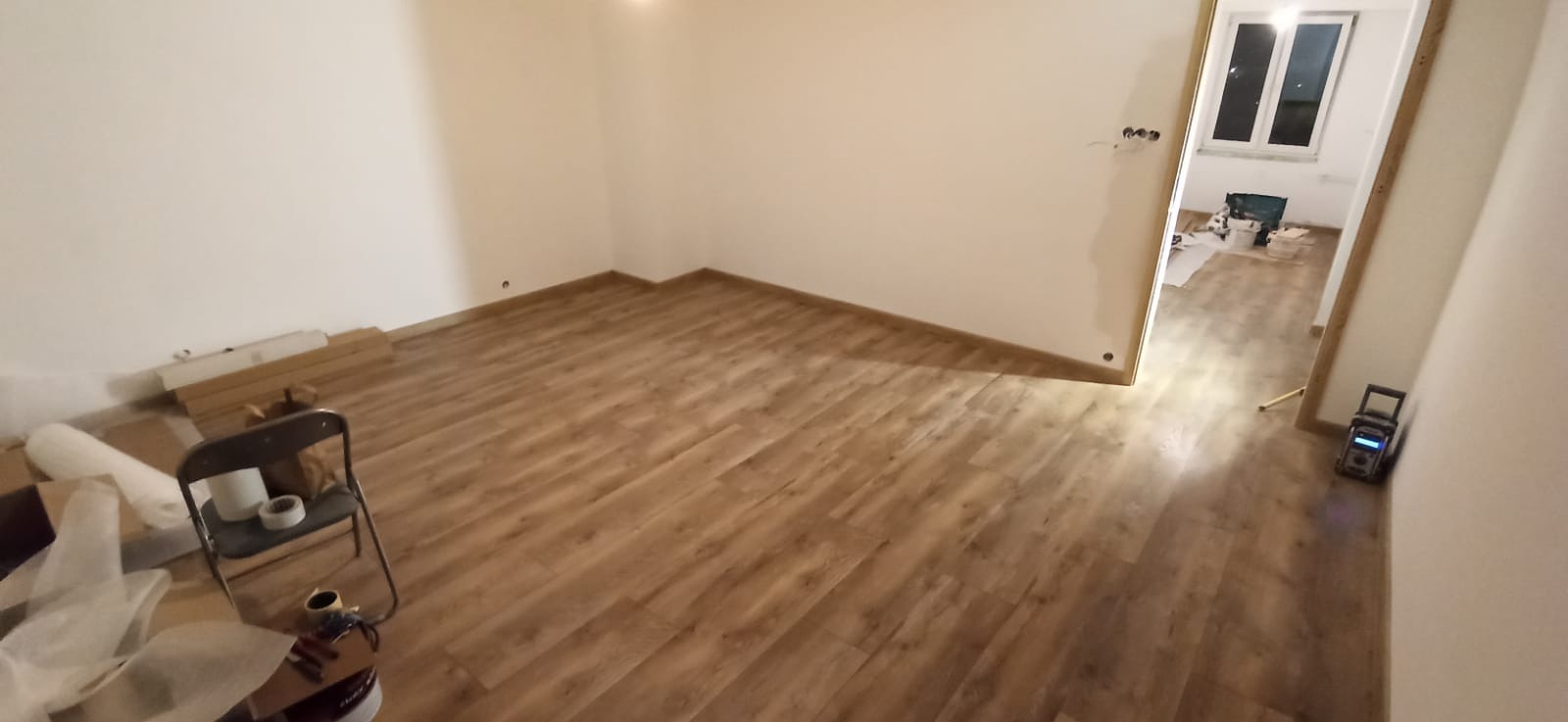 Pokój po ułożeniu paneli podłogowych w odcieniach brązu, widoczne listwy przypodłogowe, ściany przygotowane do malowania, w tle otwarte drzwi do kolejnego pomieszczenia z materiałami budowlanymi.