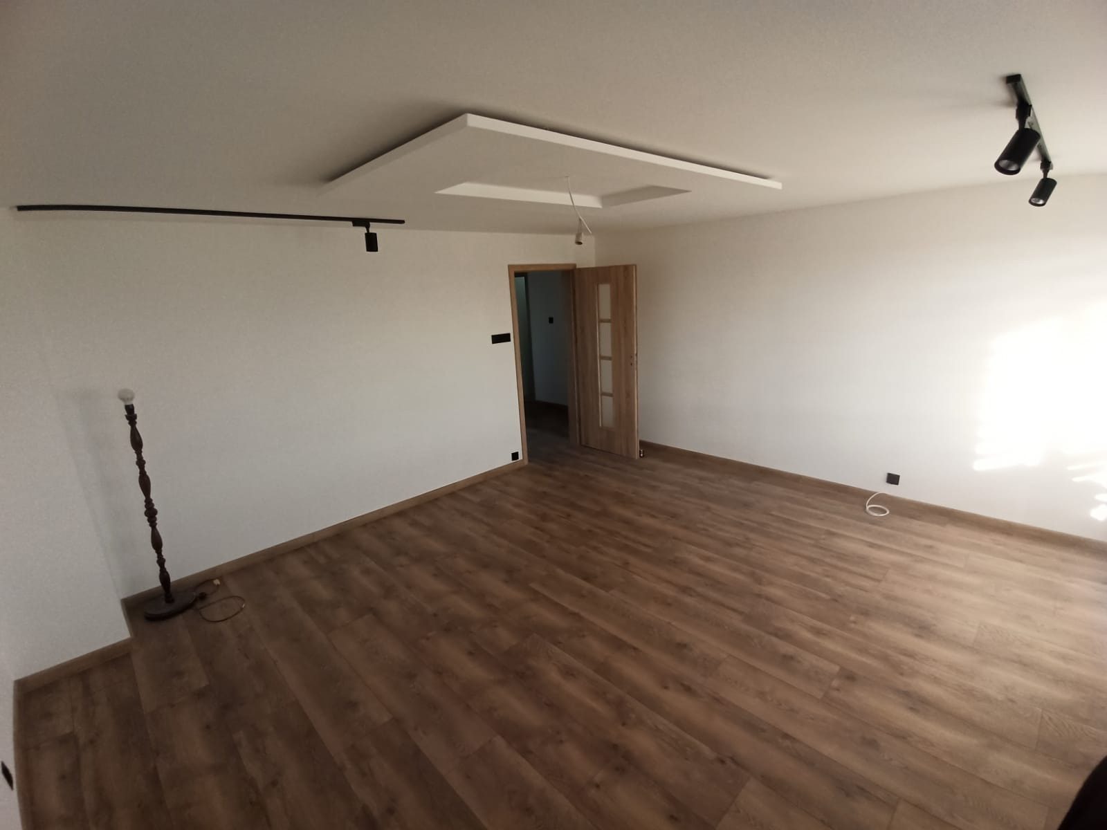 Pusty pokój po renowacji z panelami podłogowymi w odcieniach brązu, białe ściany, nowoczesne oświetlenie sufitowe w postaci reflektorów punktowych i dekoracyjnej wnęki z podwieszanym sufitem...