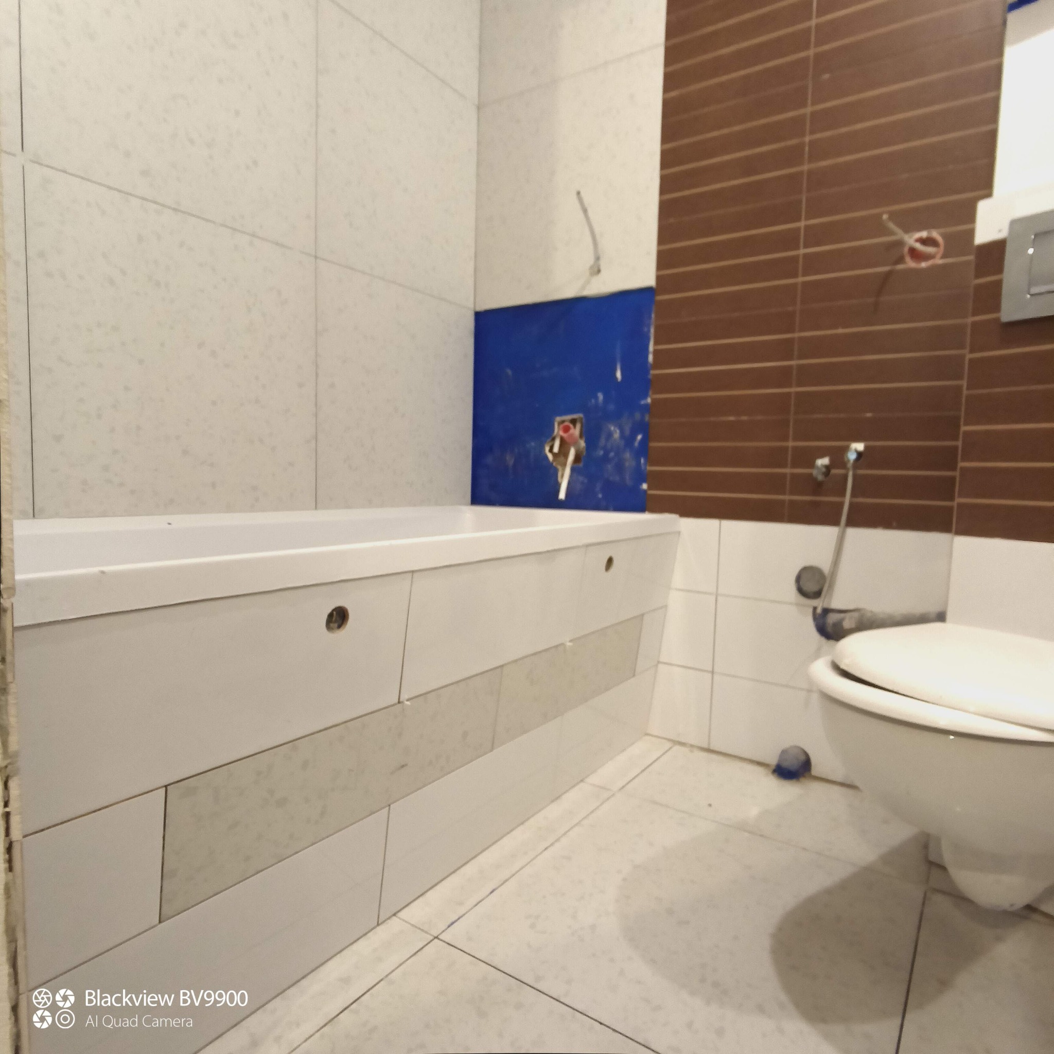 Łazienka w trakcie prac: częściowo ułożone białe i brązowe płytki na ścianach, biała wanna z widocznymi otworami, biała toaleta, widoczne instalacje hydrauliczne.