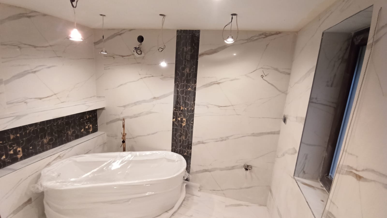 Nowa łazienka w trakcie wykańczania, z wanną owiniętą folią ochronną, marmurowe płytki na ścianach i podłodze, dekoracyjny pas z ciemnej mozaiki, widoczne niedokończone instalacje elektryczne.