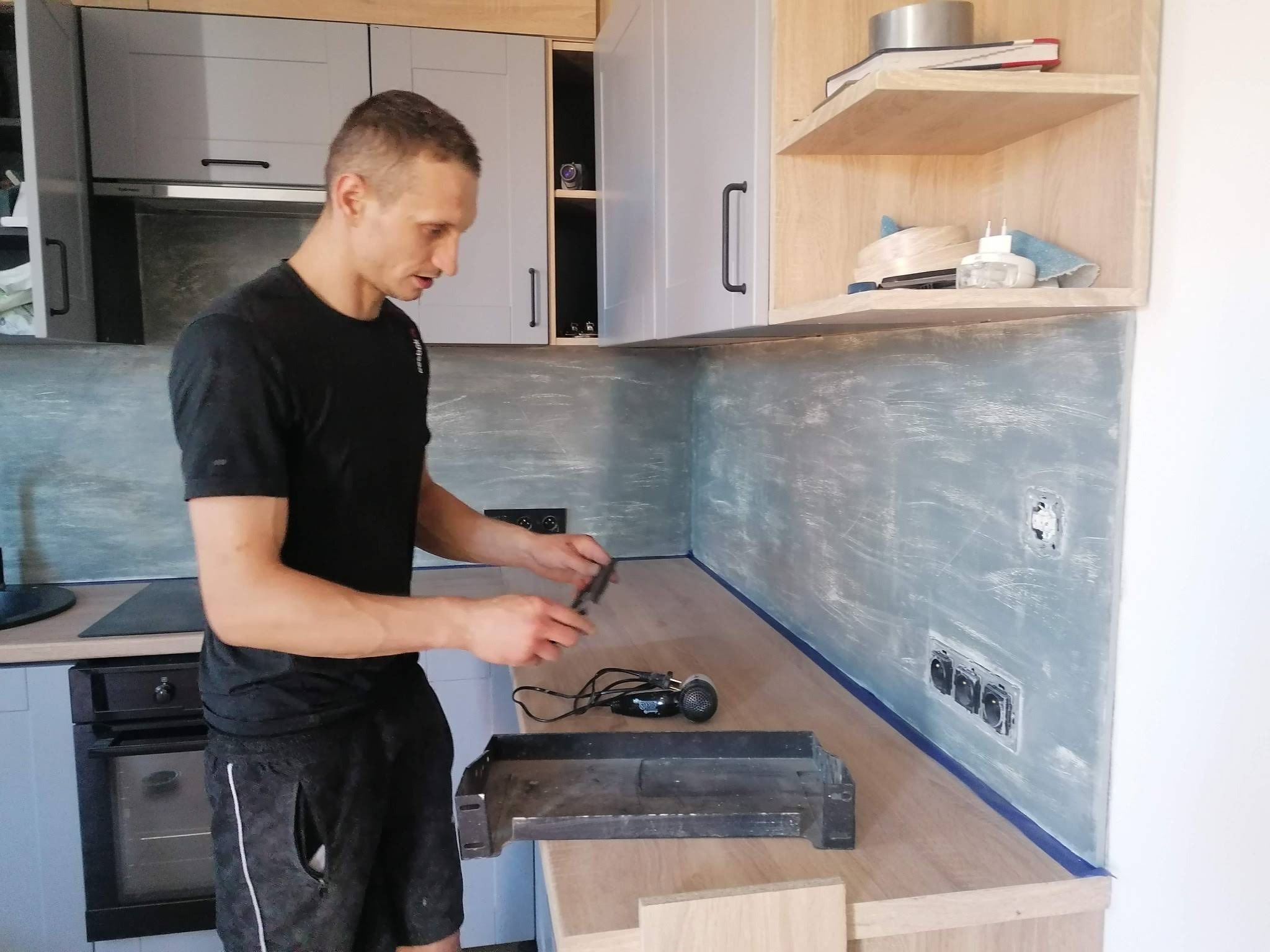 Montaż blatu kuchennego: mężczyzna przykłada listwę do blatu z jasnego drewna, ściana zabezpieczona niebieską taśmą malarską, widoczne gniazdka elektryczne i szpachlowana ściana.