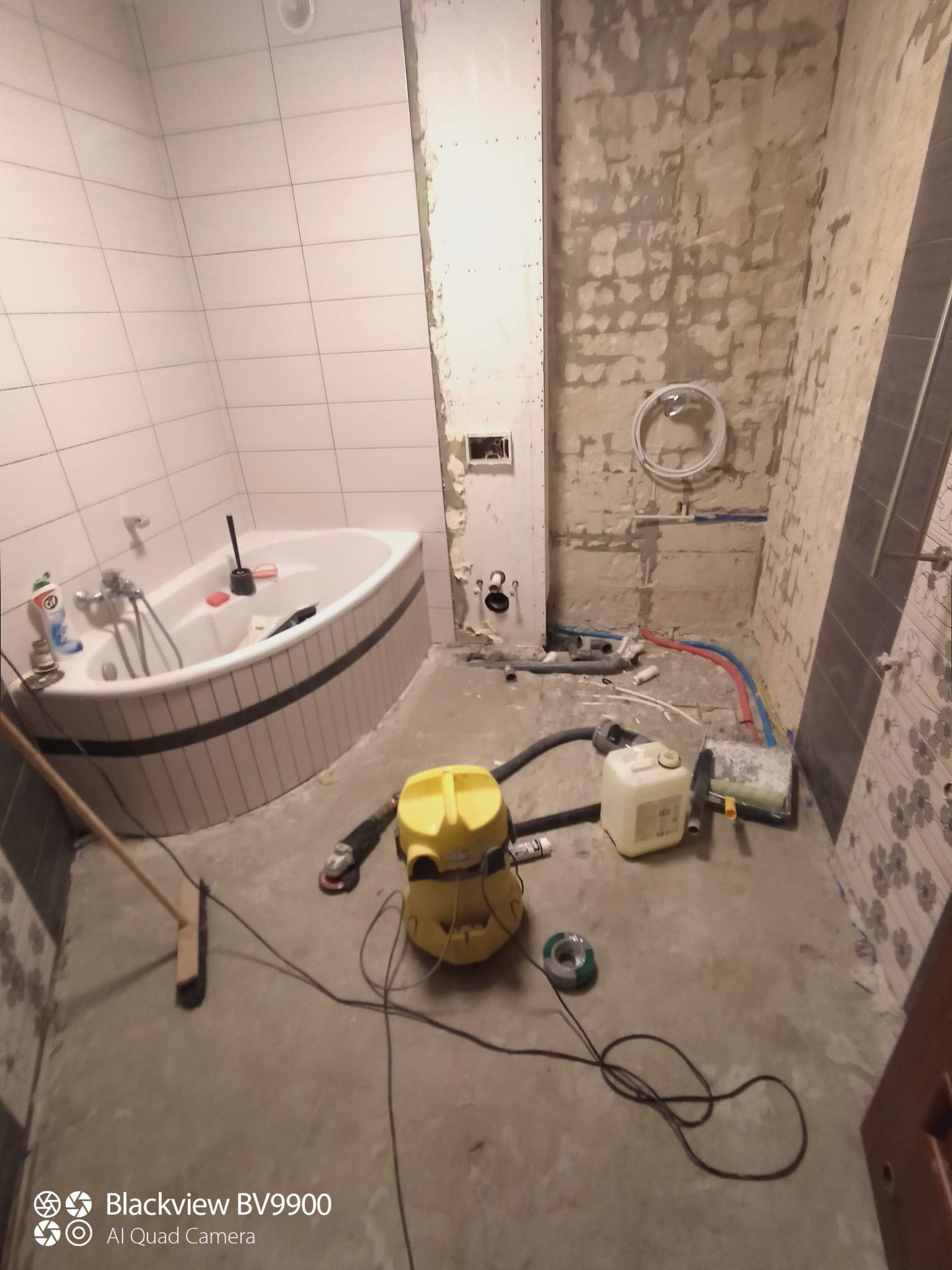 Remontowana łazienka z narożną wanną w trakcie prac: widoczne odsłonięte ściany, częściowo położone płytki, rury, żółty odkurzacz przemysłowy i narzędzia na podłodze.