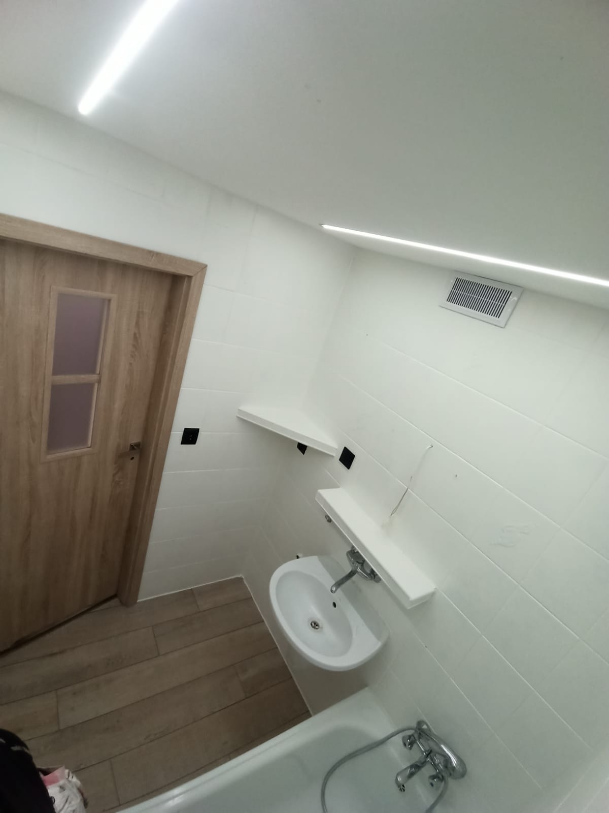 Biała łazienka z drewnianą podłogą i drzwiami, oświetlona listwami LED, z umywalką, wanną i półkami na ścianie wyłożonej białymi płytkami.