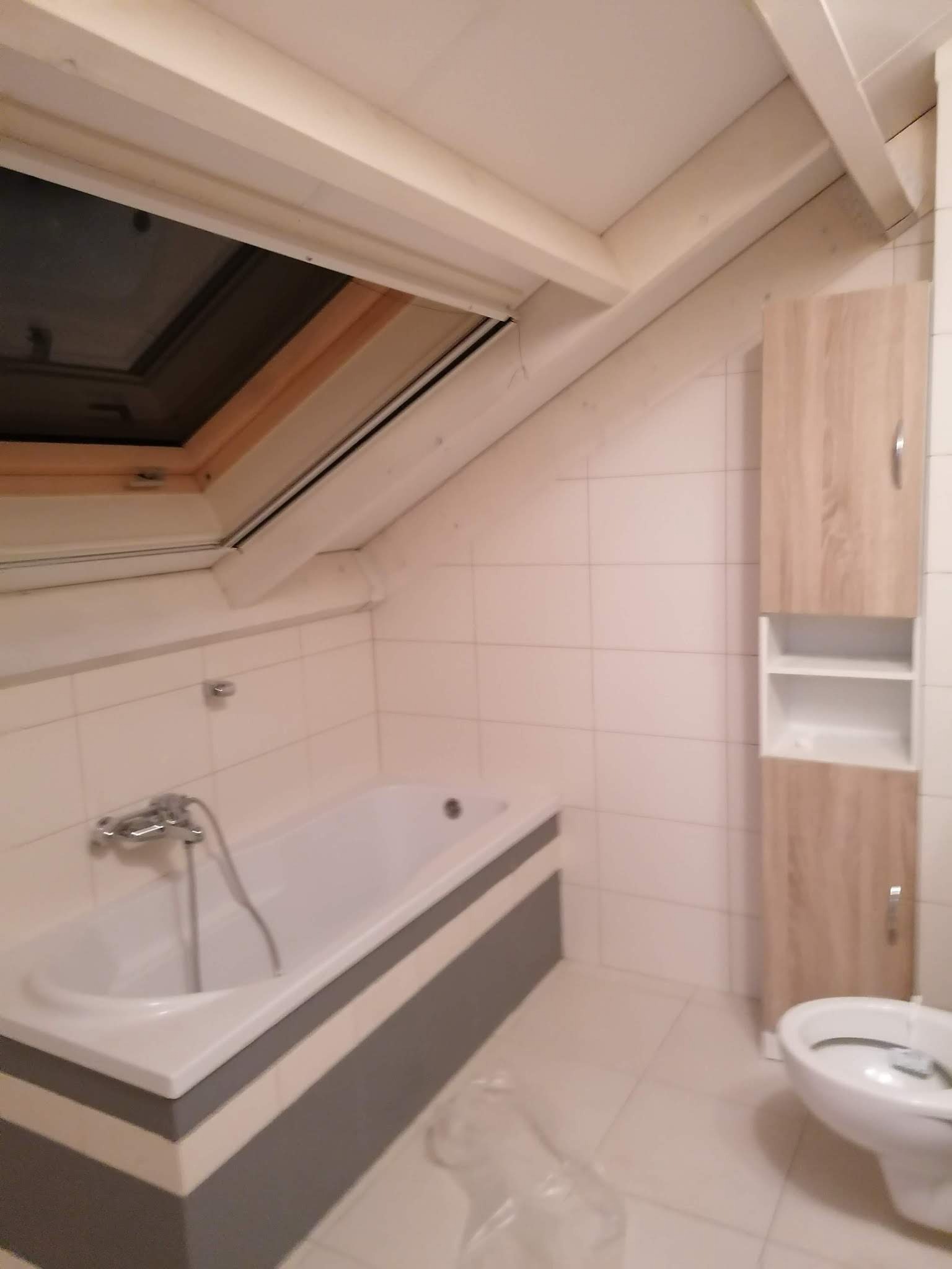 Łazienka na poddaszu z oknem dachowym, biała wanna z szarą obudową, zabudowana szafka z półkami w kolorze drewna i białe płytki na ścianach.