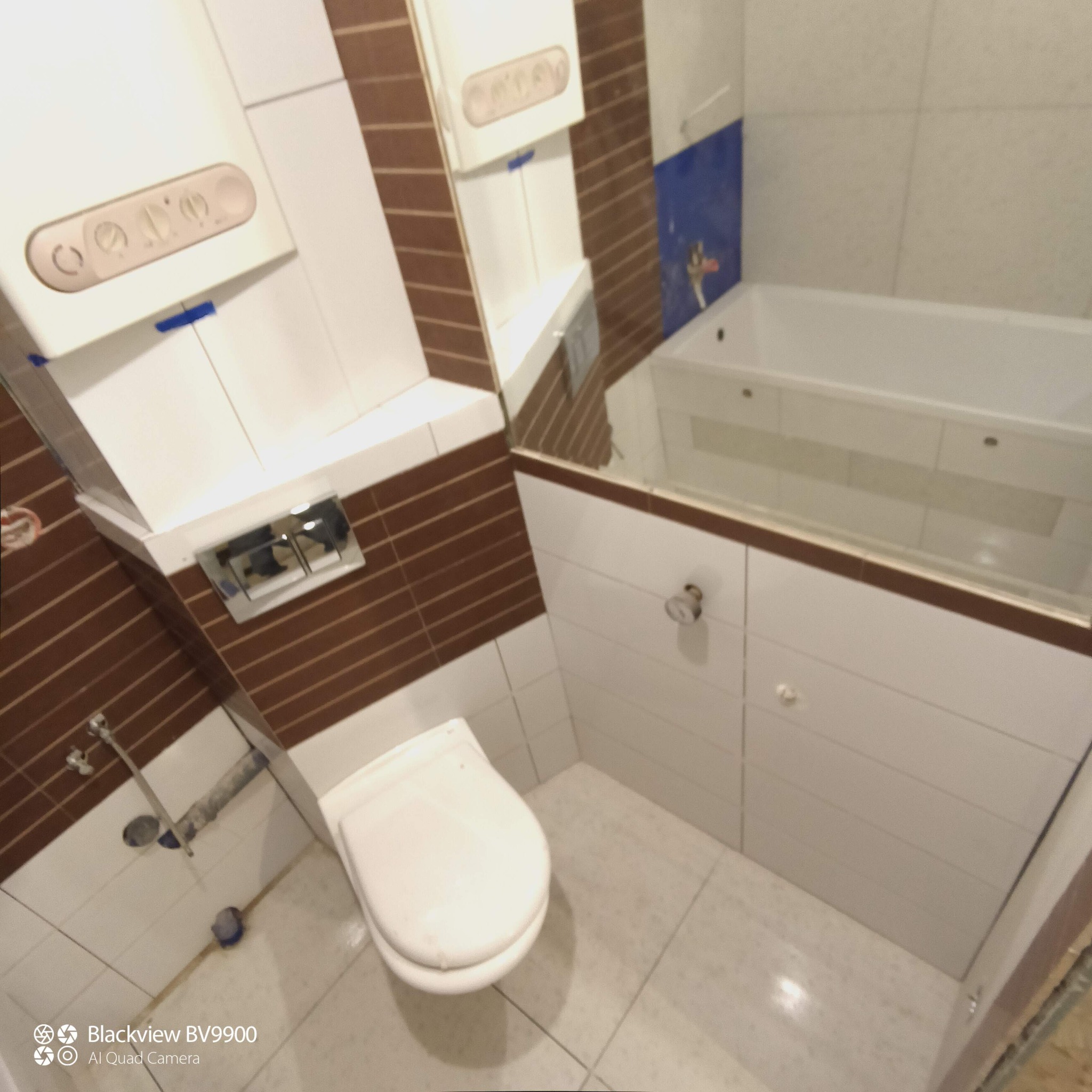 Wnętrze łazienki z brązowymi płytkami i białymi meblami, widoczny bojler, sedes i wanna z hydromasażem