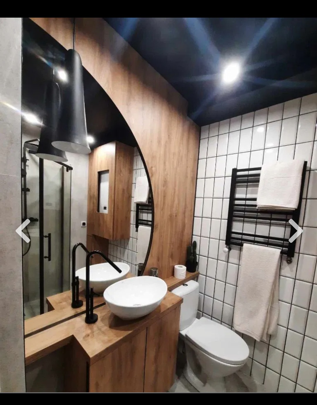 Nowoczesna łazienka z drewnianymi elementami: szafka pod umywalką z dwoma białymi umywalkami nablatowymi, czarna armatura, duże lustro z podświetleniem, białe kafelki na ścianach i czarny sufit.