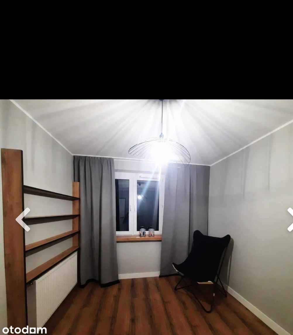 Pokój z panelami podłogowymi w odcieniach brązu, szare ściany, okno z szarymi zasłonami, drewniany regał obok grzejnika, czarne krzesło motylkowe i nowoczesna lampa wisząca.