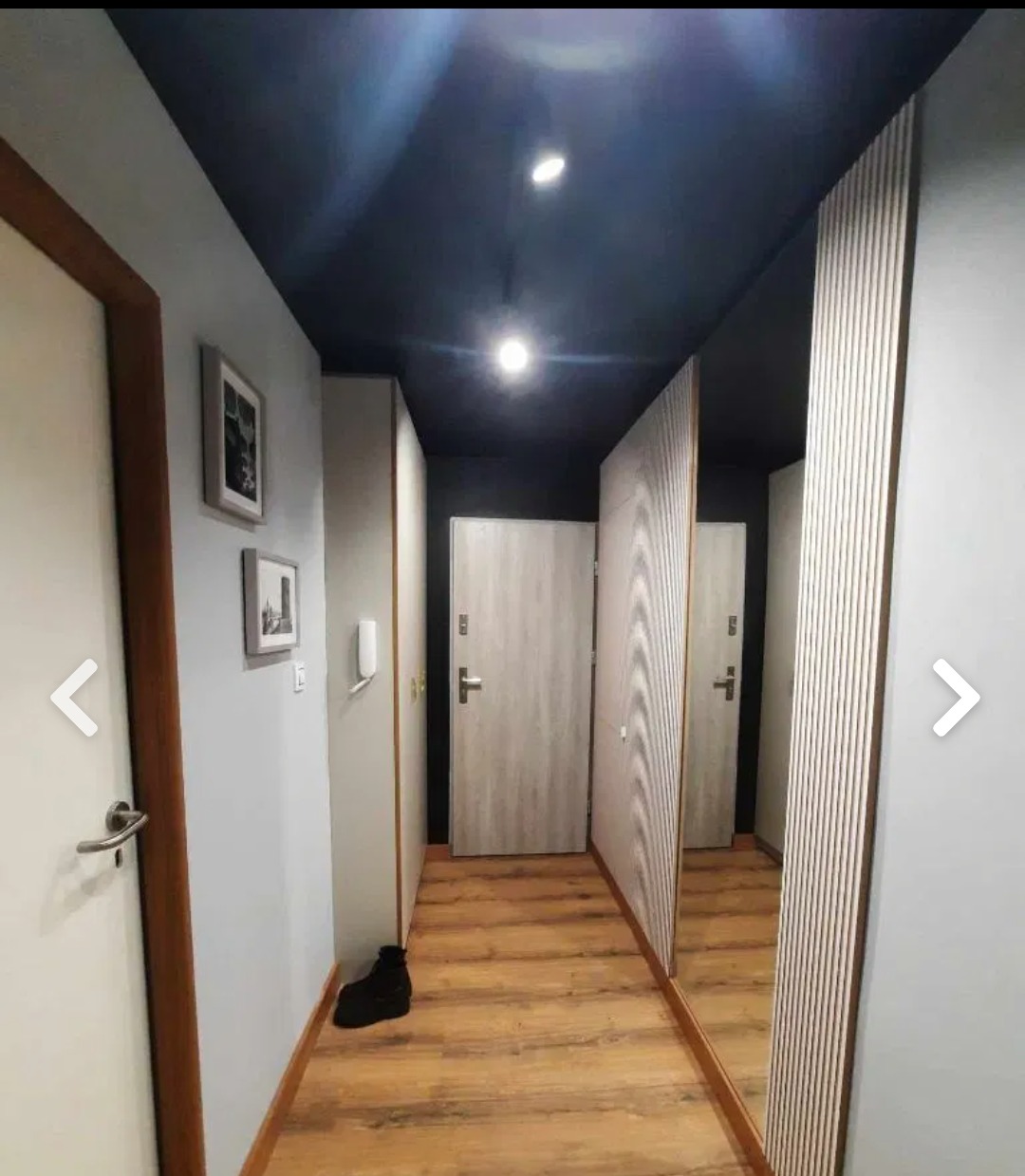 Nowoczesny przedpokój z drewnopodobnymi panelami podłogowymi, szarą ścianą z dekoracyjnymi obrazkami, ciemnym sufitem z punktowym oświetleniem i lustrem z pionowymi listwami.