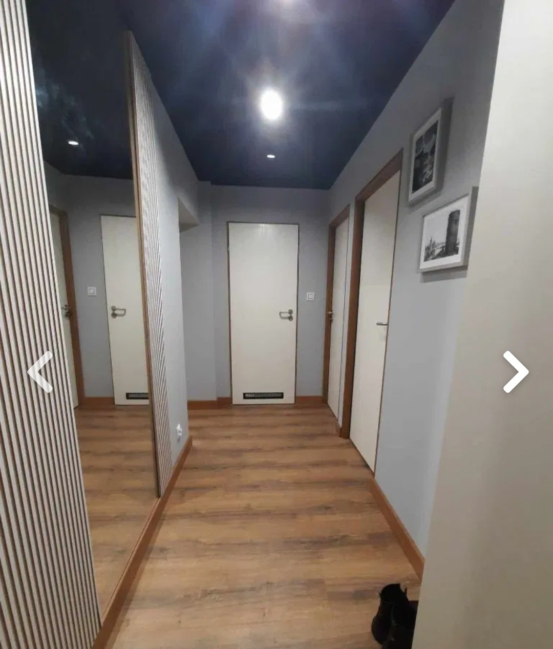 Korytarz po renowacji z panelami podłogowymi w odcieniach brązu, szarymi ścianami, ciemnym sufitem z oświetleniem punktowym, białymi drzwiami i dekoracyjnymi obrazami na ścianie.