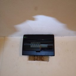 INSTALTEAM-ŚWIDNICA - Czarna skrzynka rozdzielcza elektryczna marki ELEQANT zamontowana na ścianie z widocznym wycięciem w tynku, sufit z nierówną fakturą.