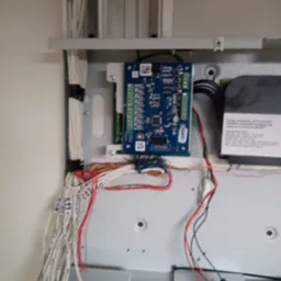 Płyta sterująca z podłączonymi przewodami wewnątrz metalowej obudowy, część przewodów oznaczona etykietami, widoczna naklejka ostrzegawcza na obudowie.