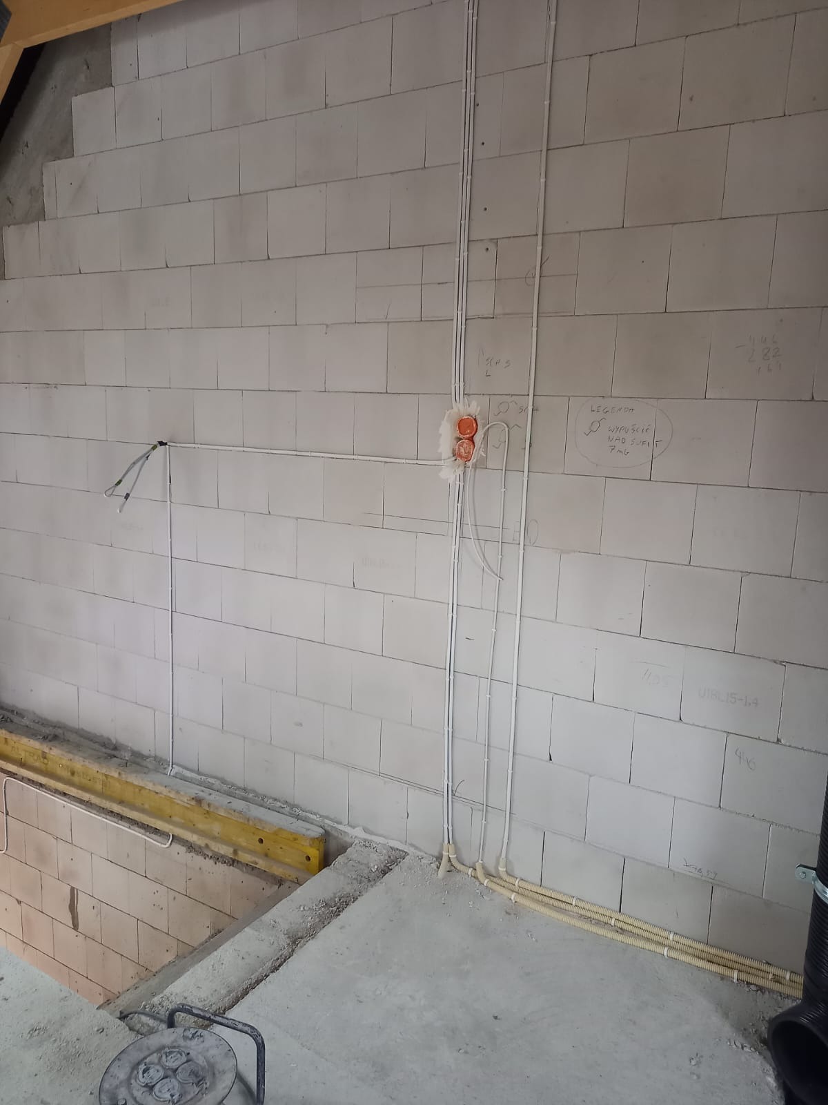 Wewnętrzna instalacja elektryczna w trakcie montażu w surowym budynku z białego betonu komórkowego, widoczne kable w peszlach i puszki instalacyjne, na ścianie odręczne notatki.