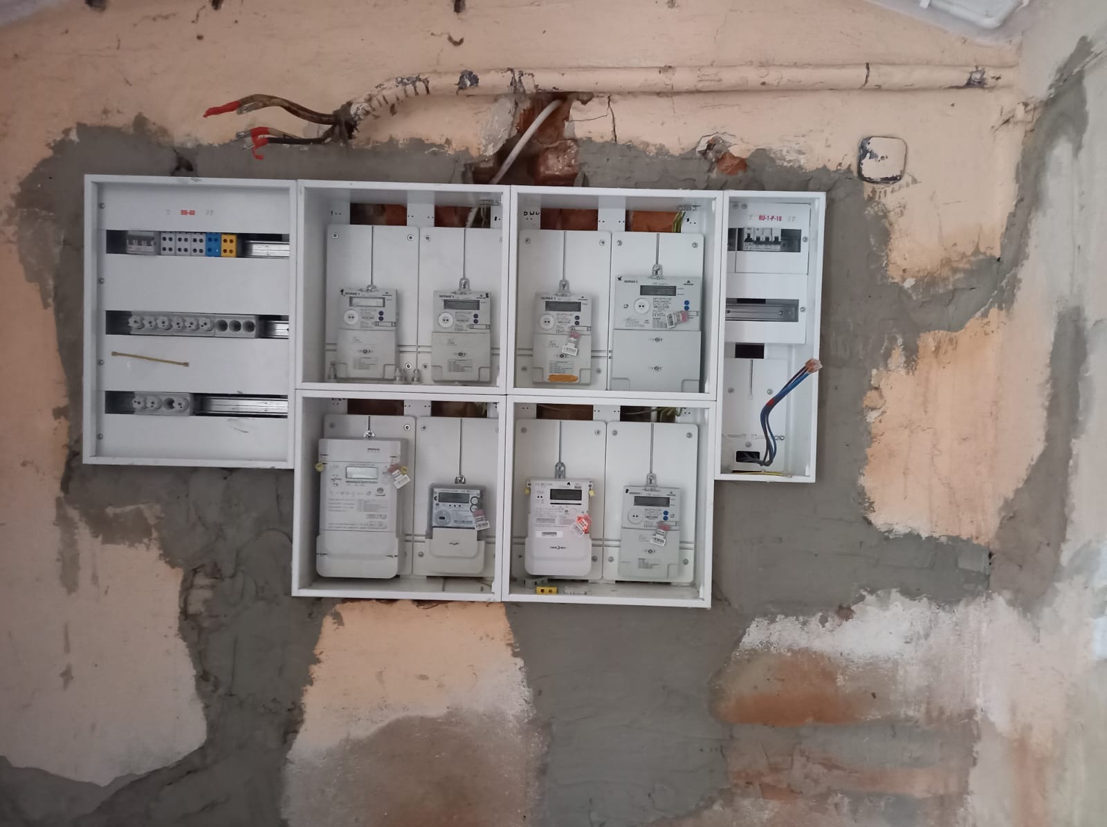 Szafka rozdzielcza z licznikami energii elektrycznej zamontowana na surowej ścianie w trakcie wykończenia, widoczne przewody elektryczne i rury instalacyjne.