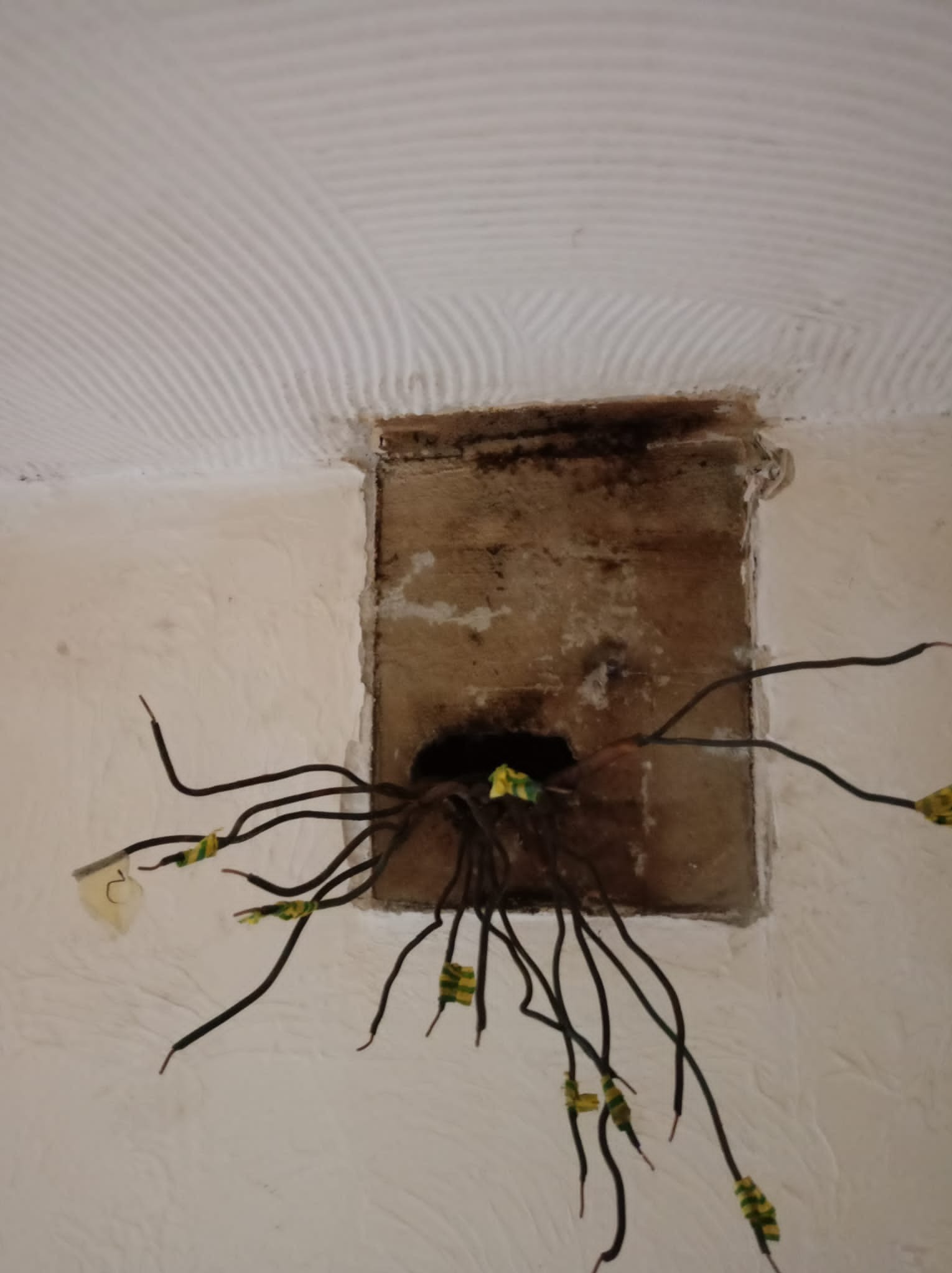 Otwór w ścianie z wystającymi kablami elektrycznymi zabezpieczonymi taśmą izolacyjną w kolorze żółto-zielonym, widoczne ślady po instalacji i nierówna powierzchnia ściany.