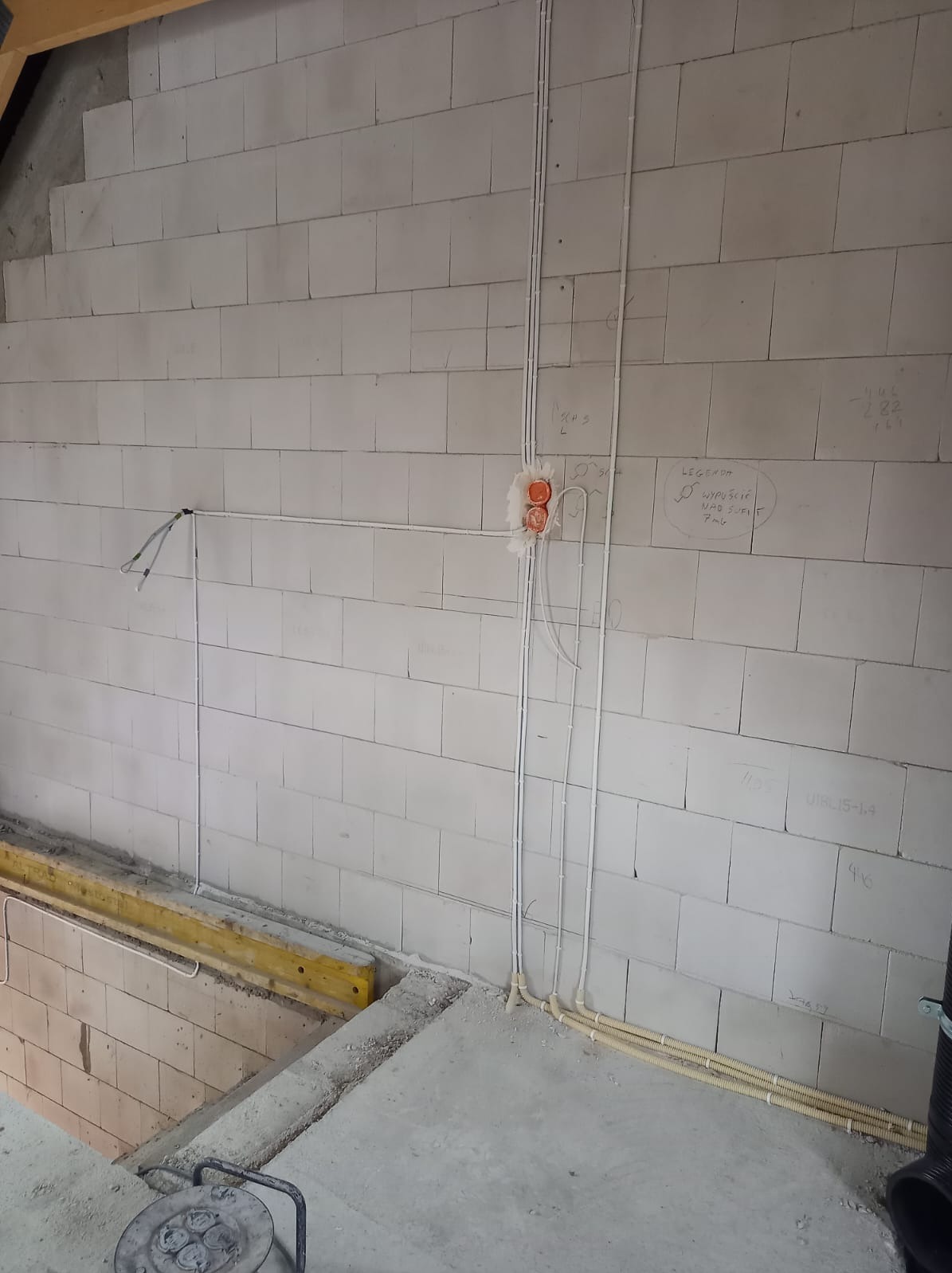 Instalacja elektryczna w trakcie montażu w surowym wnętrzu z białych bloczków, widoczne przewody w peszlach i puszki elektryczne, na ścianie odręczne notatki.