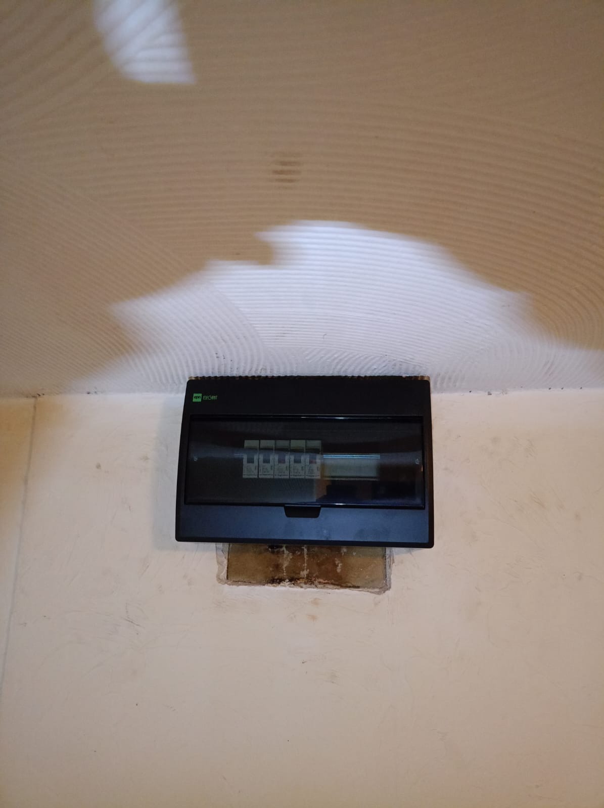 Czarna skrzynka rozdzielcza elektryczna marki ELEQANT zamontowana na ścianie z widocznym wycięciem w tynku, sufit z nierówną fakturą.