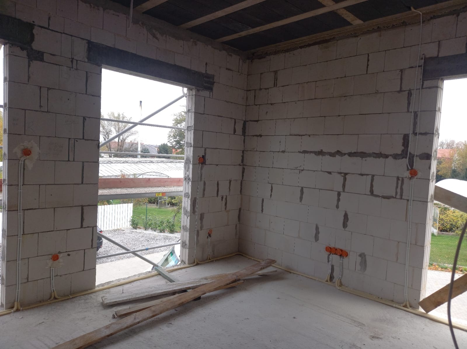 Surowy stan budynku w trakcie prac: widoczne ściany z bloczków, rozprowadzone przewody elektryczne w peszlach, puszki instalacyjne w kolorze pomarańczowym, fragment rusztowania za oknem.