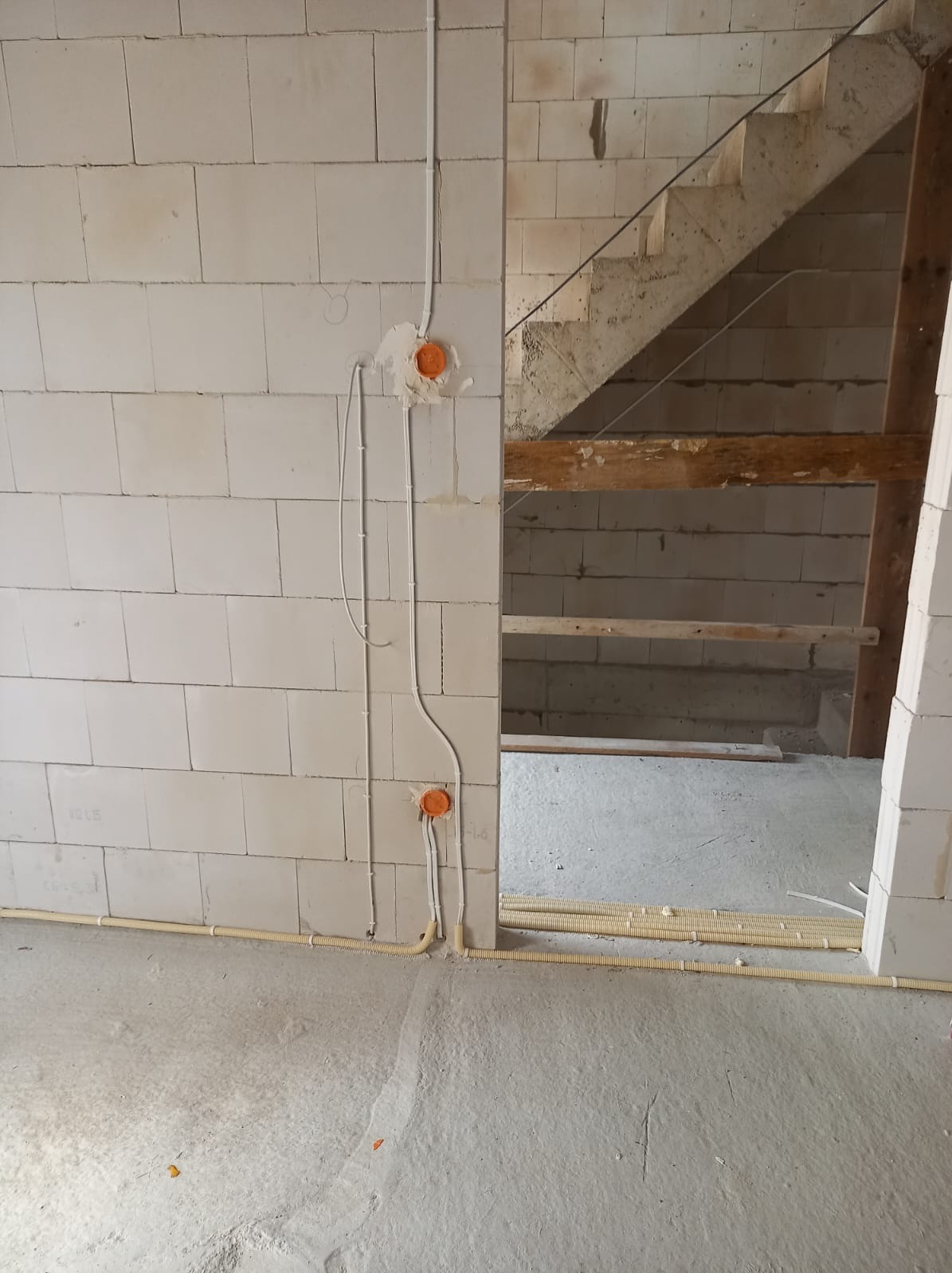 Surowy stan: Kable elektryczne w peszlach i puszki montażowe na ścianie z bloczków betonowych, widoczne schody w tle.