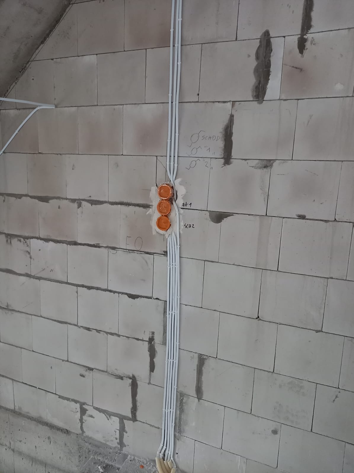 Surowe ściany z betonu komórkowego z trzema pomarańczowymi puszkami podtynkowymi i pionowo ułożonymi przewodami elektrycznymi w białych peszlach, z widocznymi odręcznymi notatkami na ścianie.