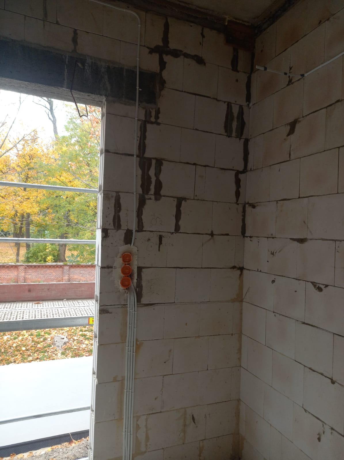 Surowa ściana z betonu komórkowego w trakcie budowy, widoczne pomarańczowe puszki elektryczne i białe przewody prowadzone po ścianie, na zewnątrz widok na drzewa w jesiennych barwach i fragment...