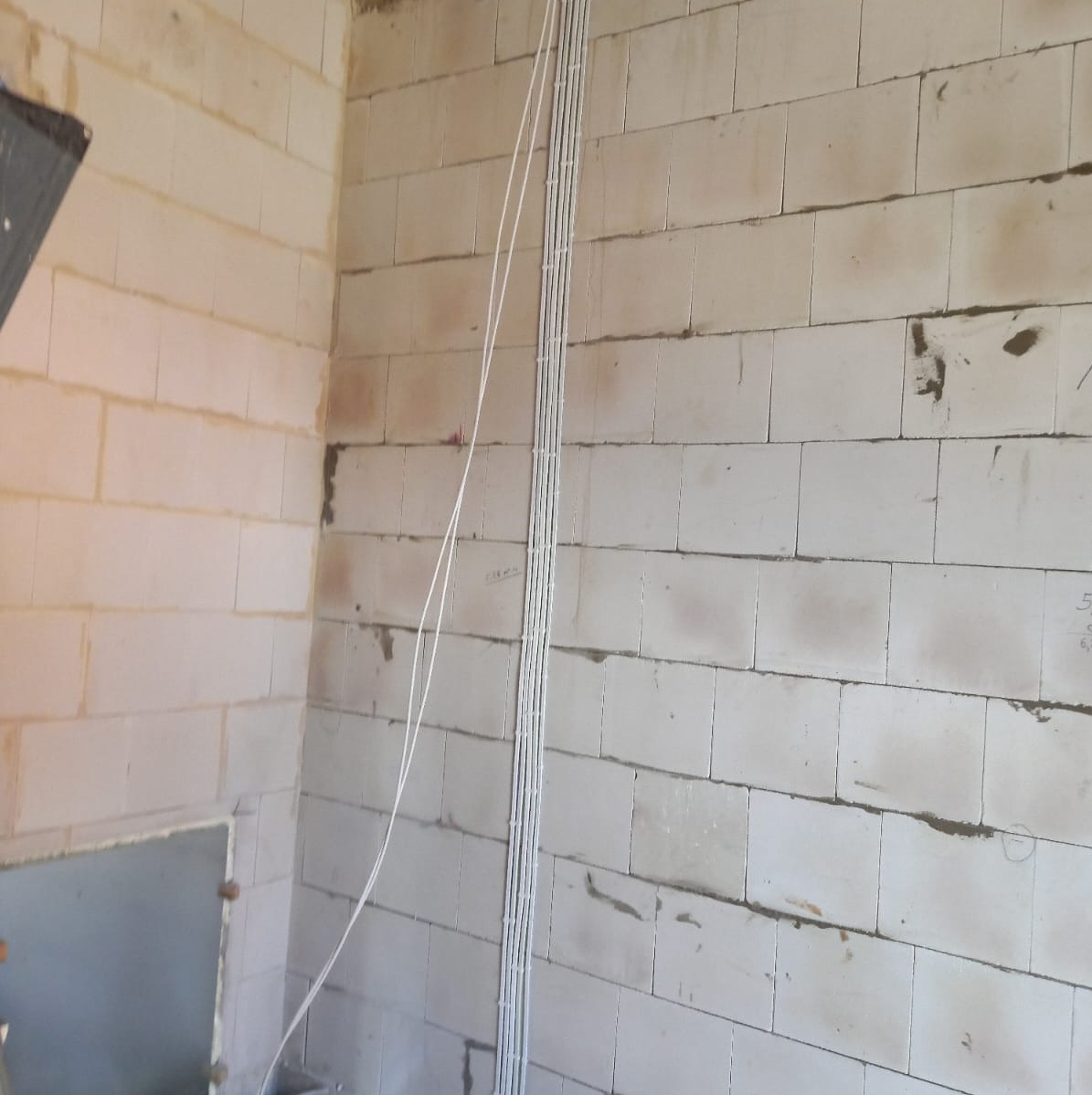 Białe przewody elektryczne prowadzone pionowo po ścianie z bloczków betonowych, widoczne ślady zaprawy.