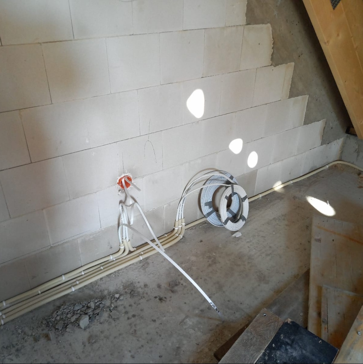 Surowy stan poddasza z widoczną instalacją elektryczną w peszlach, puszką instalacyjną w ścianie z betonu komórkowego i zwiniętym kablem na podłodze.