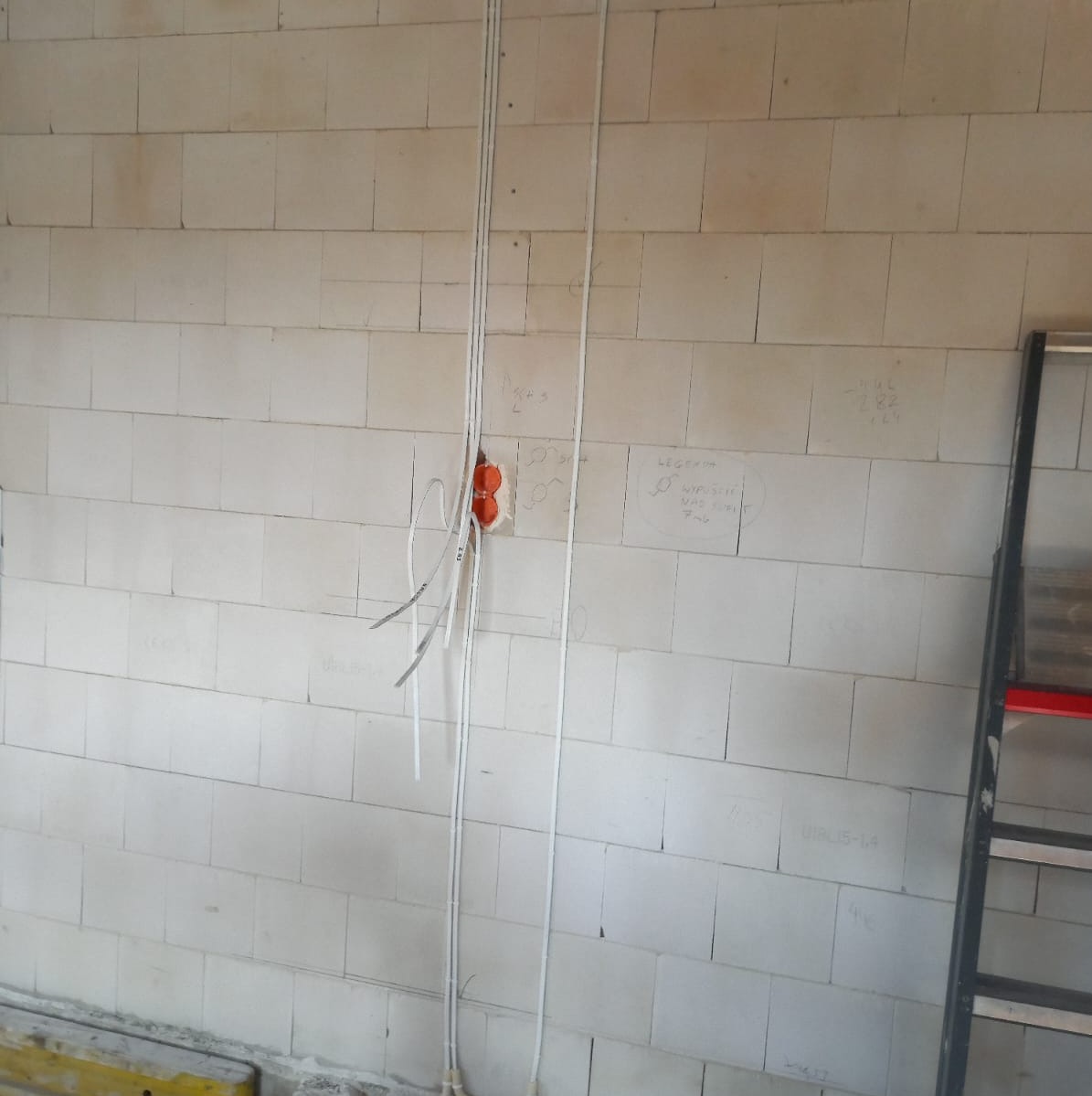 Ściana z betonu komórkowego w Świdnicy z widocznymi przewodami elektrycznymi w peszlach i puszkami instalacyjnymi, obok drabina, na ścianie odręczne notatki.