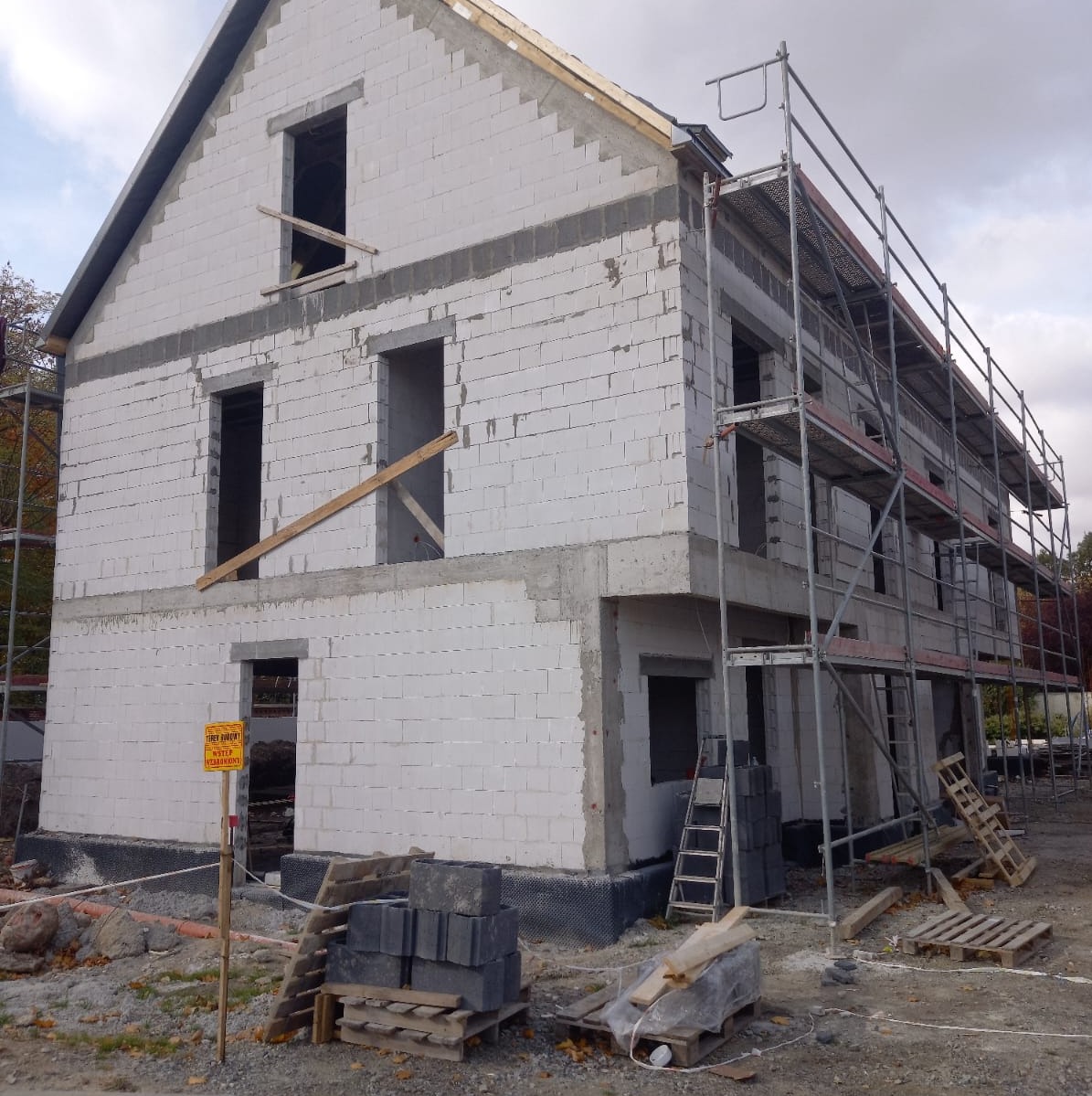 Surowy stan dwupiętrowego domu z białego bloczka, widoczne rusztowania i przygotowane materiały budowlane, tabliczka ostrzegawcza na pierwszym planie.