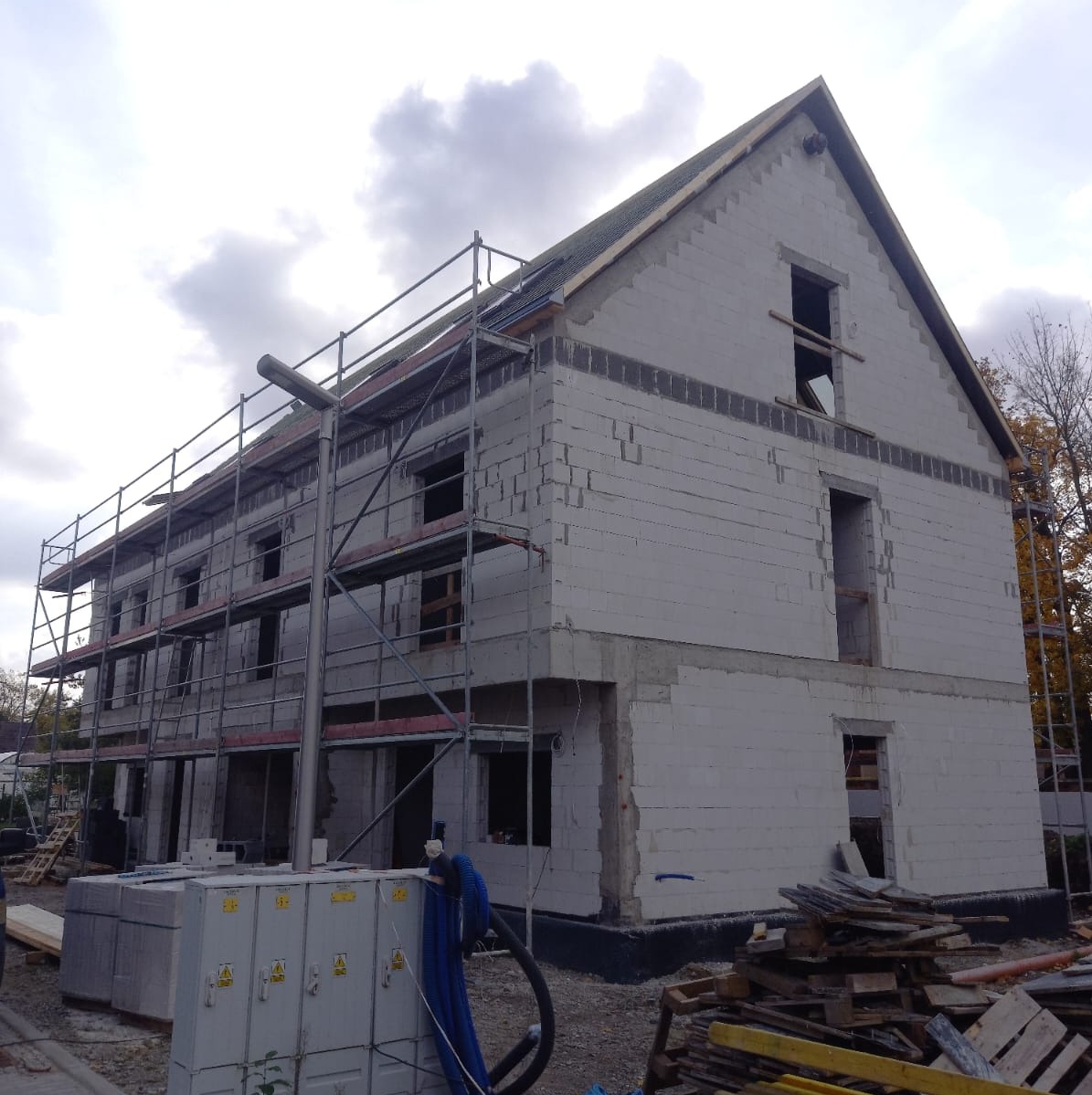 Surowy stan dwupiętrowego budynku z białych bloczków, z otworami okiennymi i drzwiami, otoczonego rusztowaniami, z widocznymi materiałami budowlanymi i skrzynką elektryczną na pierwszym planie.