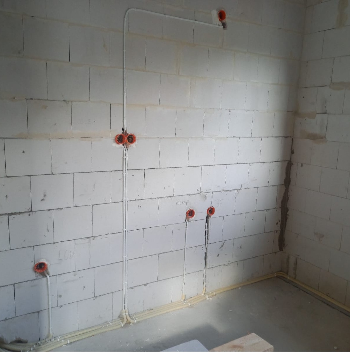 Surowy mur z betonu komórkowego z widocznymi, częściowo zamontowanymi przewodami elektrycznymi w białych rurkach i pomarańczowymi puszkami instalacyjnymi.